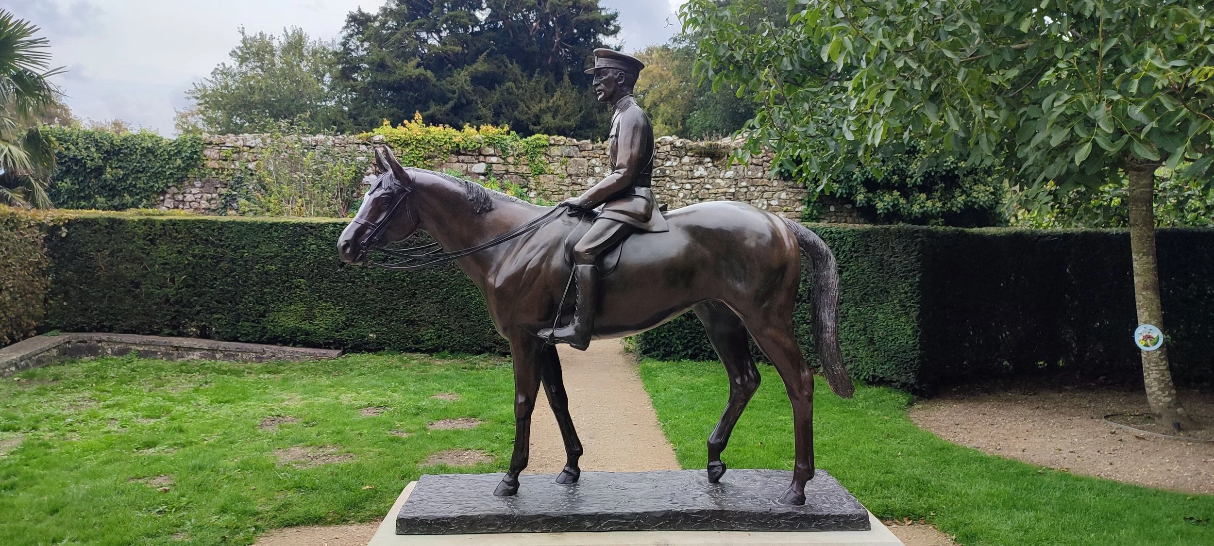 Bronze statue of a man in military uniform riding a horse, located outdoors in a park with green grass, trees, and a stone wall in the background.