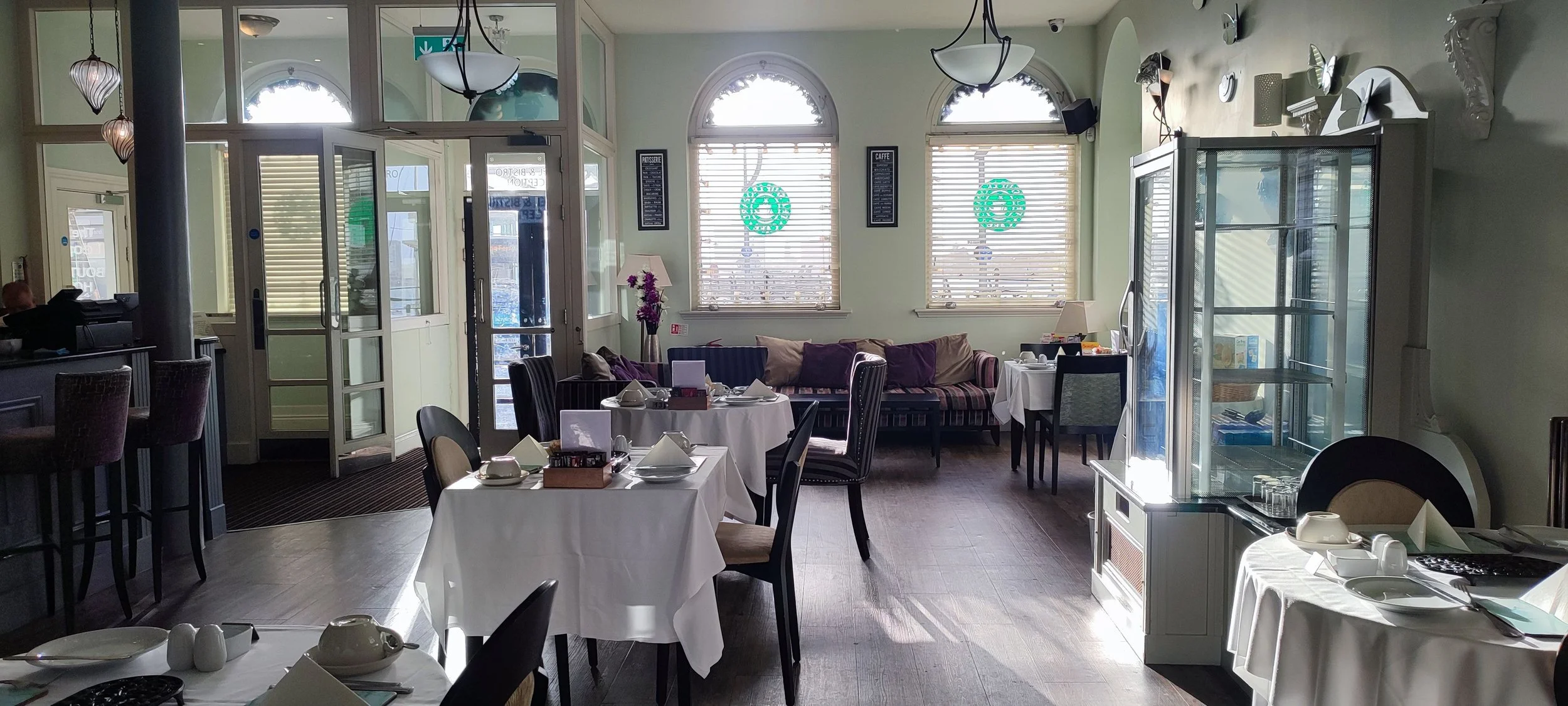 Interior of a restaurant with tables set for dining, including white tablecloths, plates, cups, and utensils. There's a sofa with cushions against the back wall and large windows with blinds letting in natural light. Decor includes flower arrangements, lamps, and wall art.