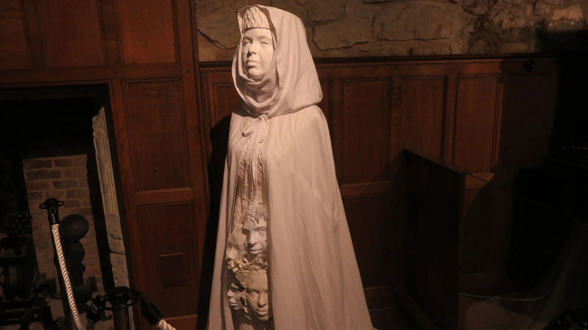 A sculpture of a woman with a hood, draped in fabric, standing in front of a wooden wall with stone accents.