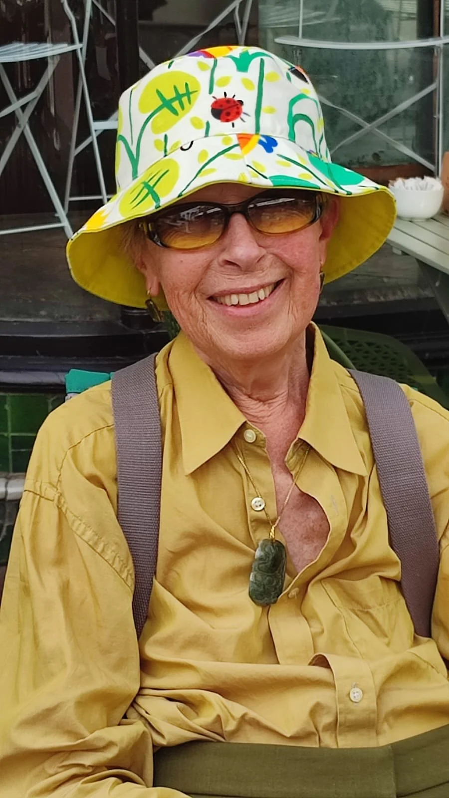 An elderly woman wearing a yellow shirt, a colorful bucket hat with a ladybug, colorful leaves, and grass pattern, sunglasses, and a necklace with a stone pendant smiling outdoors.