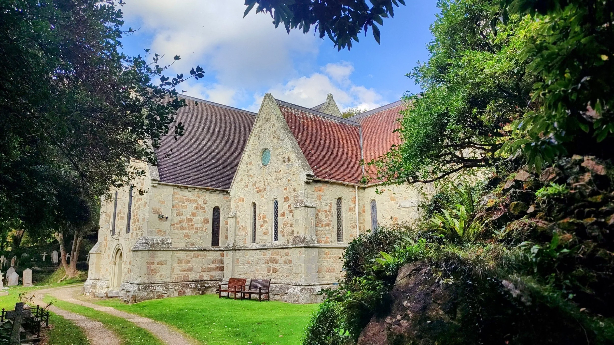 A stone church with red-tile roof, narrow windows, and circular window, surrounded by green trees, benches, and a well-kept grassy yard, under a partly cloudy sky.