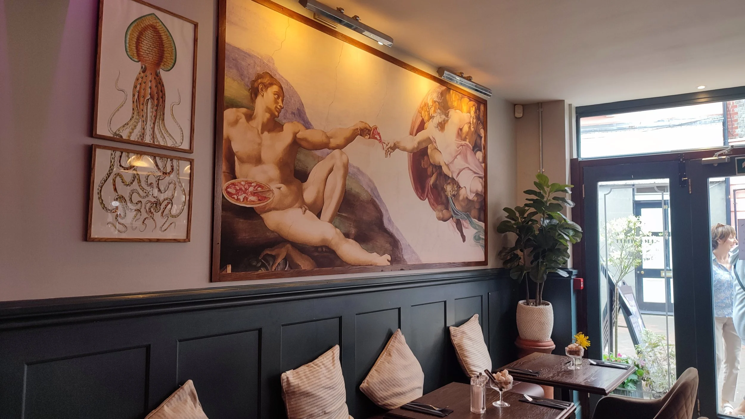 Interior of a restaurant with framed artwork on the wall, including a large Neo-Renaissance style painting of Michelangelo's Creation of Adam and two botanical illustrations of a jellyfish, with a black wainscot, beige pillows on chairs, potted plant, and people near the entrance.