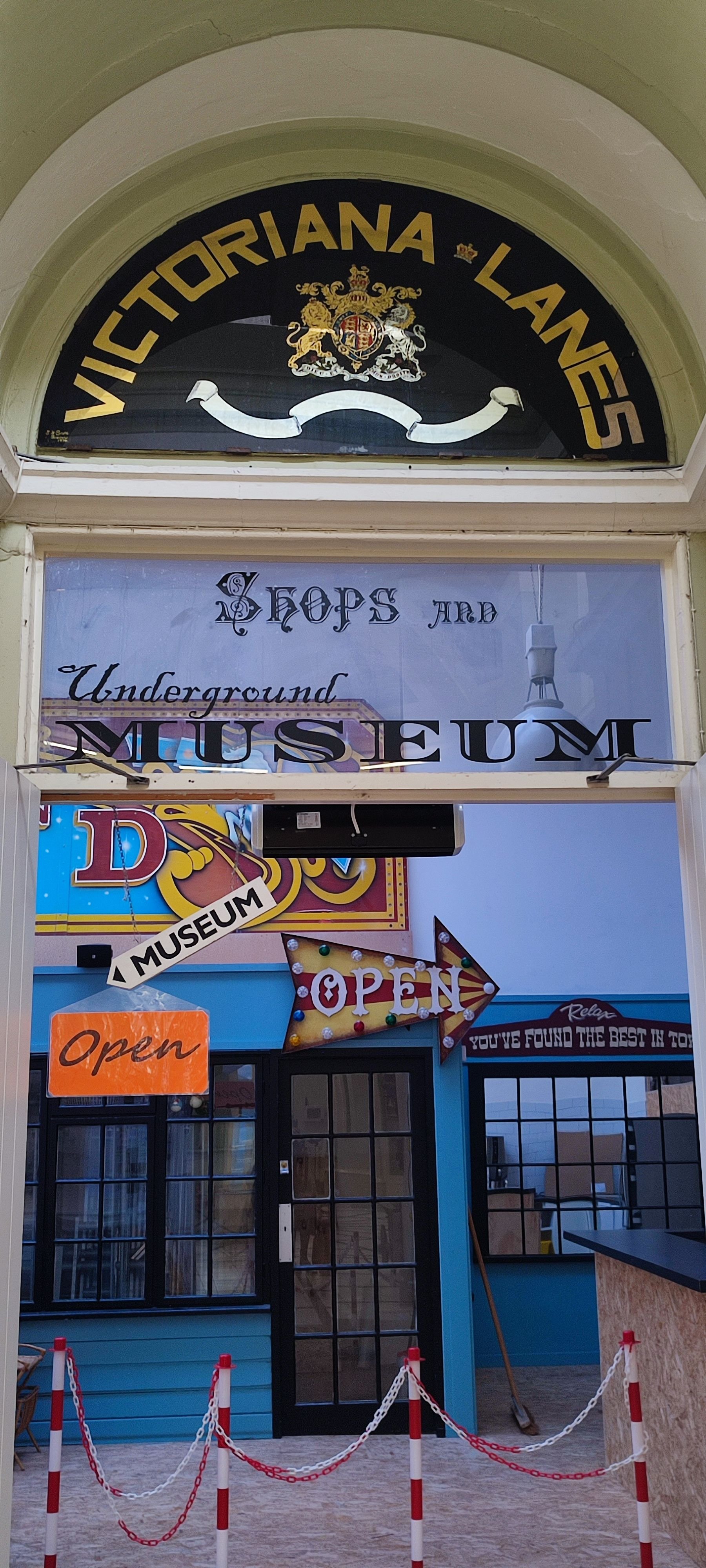Entrance to the Victoria Lanes bowling alley and underground museum. The signs indicate it is open, with a colorful arrow sign pointing inside, and there are barriers in front of the door.