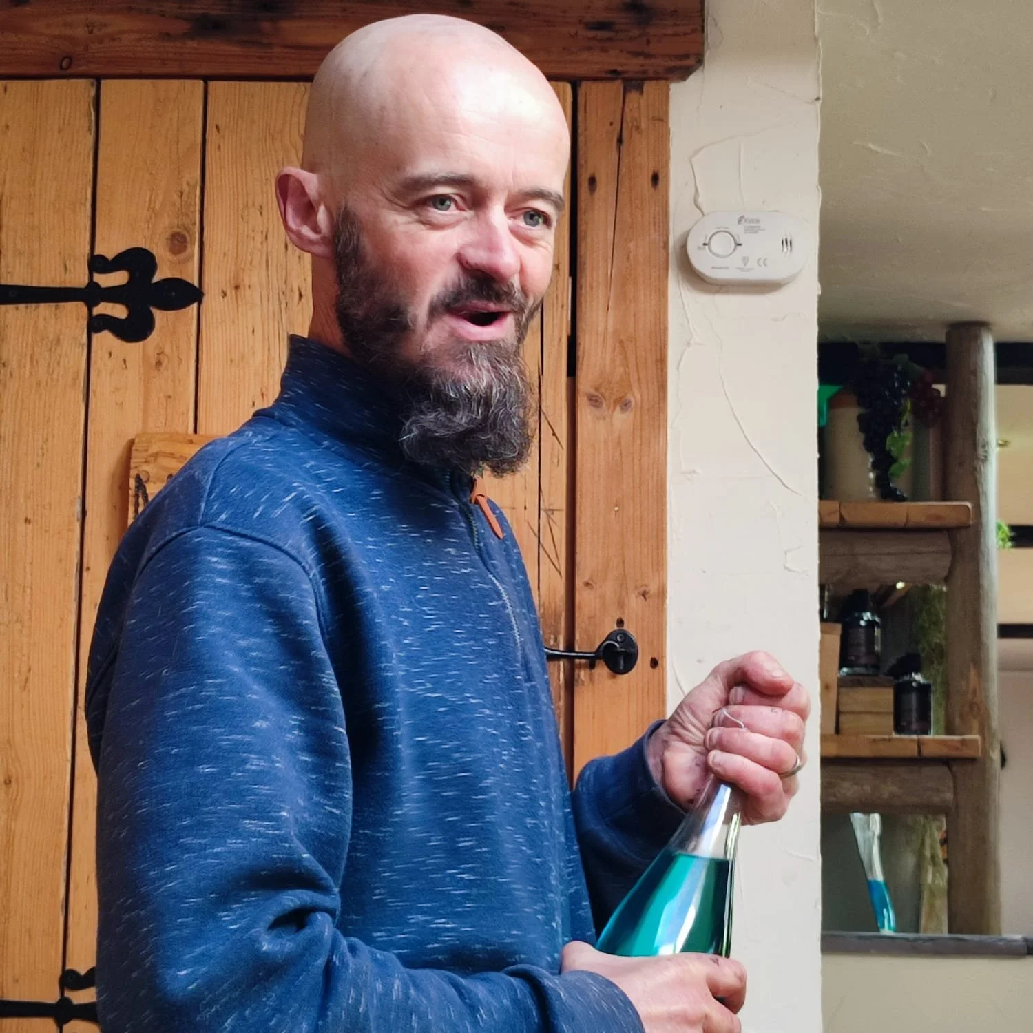 A man with a bald head and beard, wearing a blue zip-up hoodie, is holding a flask containing a blue liquid, standing indoors with wood-paneled walls and shelves in the background.