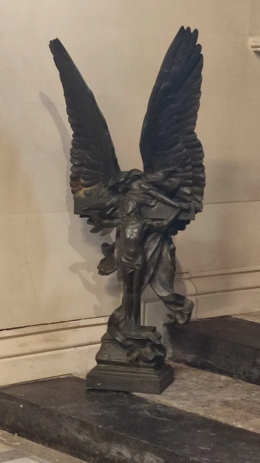 Bronze statue of a winged figure carrying a crossed flag, standing on a pedestal in front of a beige wall.