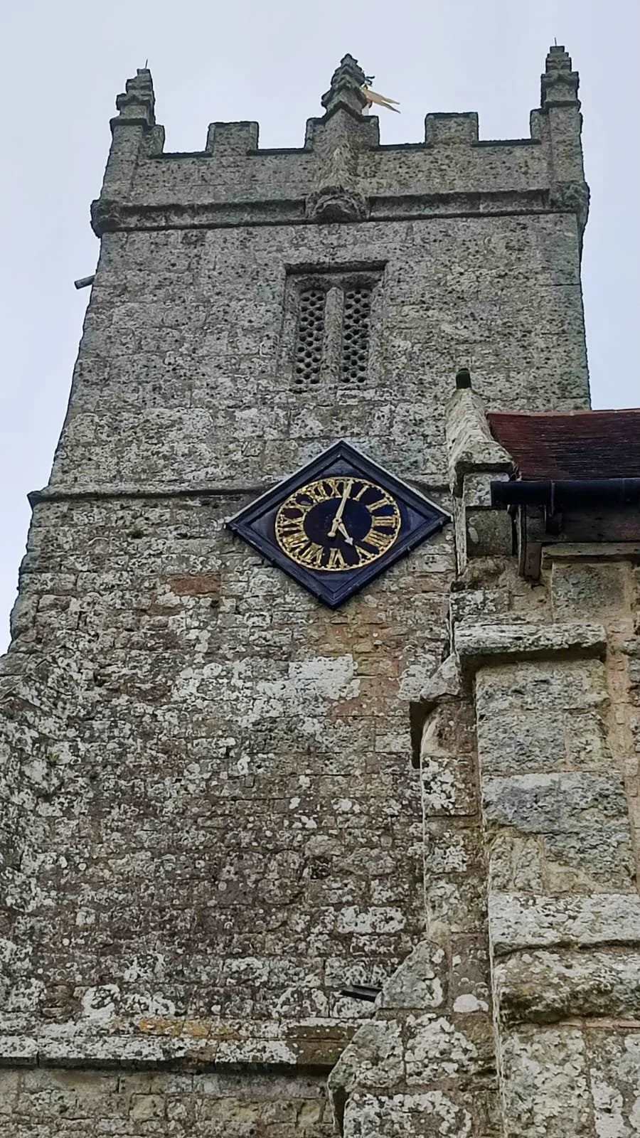 A stone church tower with a clock showing 12:02, a crenelated battlement at the top, and a weather vane at the pinnacle.