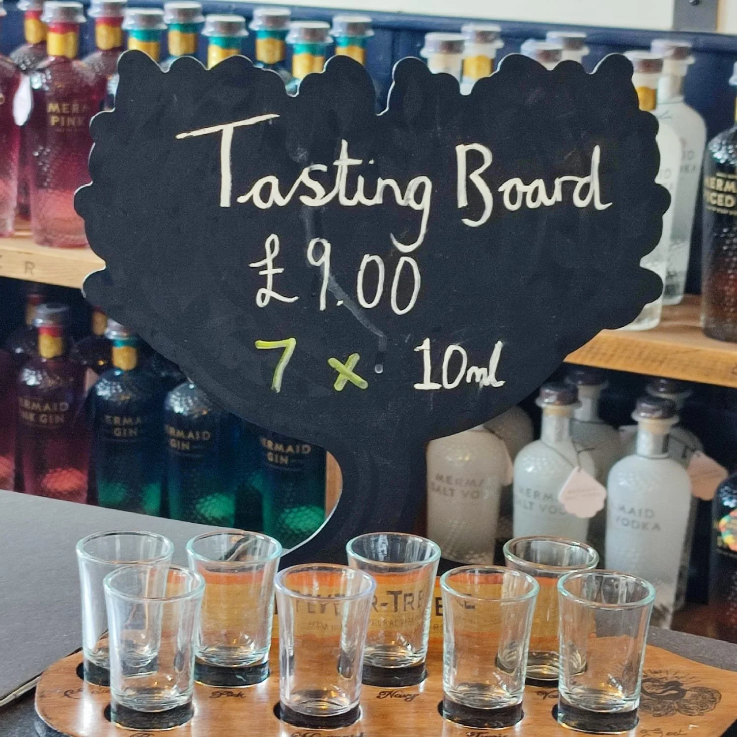 A tasting board with eight small glasses filled with small pours of spirits, set on a wooden tray in front of a chalkboard sign. The sign reads 'Tasting Board £9.00 7 x 10ml' and is black with white and yellow writing, with bottles of various spirits and tonic water in the background.