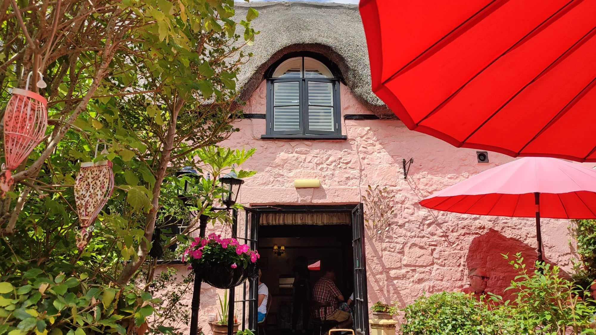 Outdoor patio with pink stone building, colorful umbrellas, lush green plants, pink flowers, and people sitting outside.