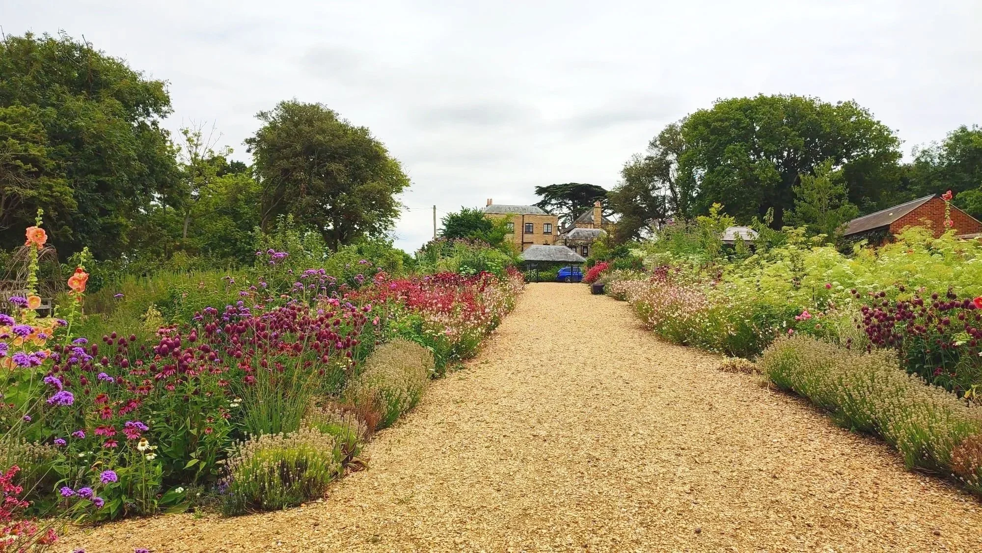 A gravel pathway flanked by colorful blooming flowers leading to a house in the background, surrounded by green trees under a cloudy sky.