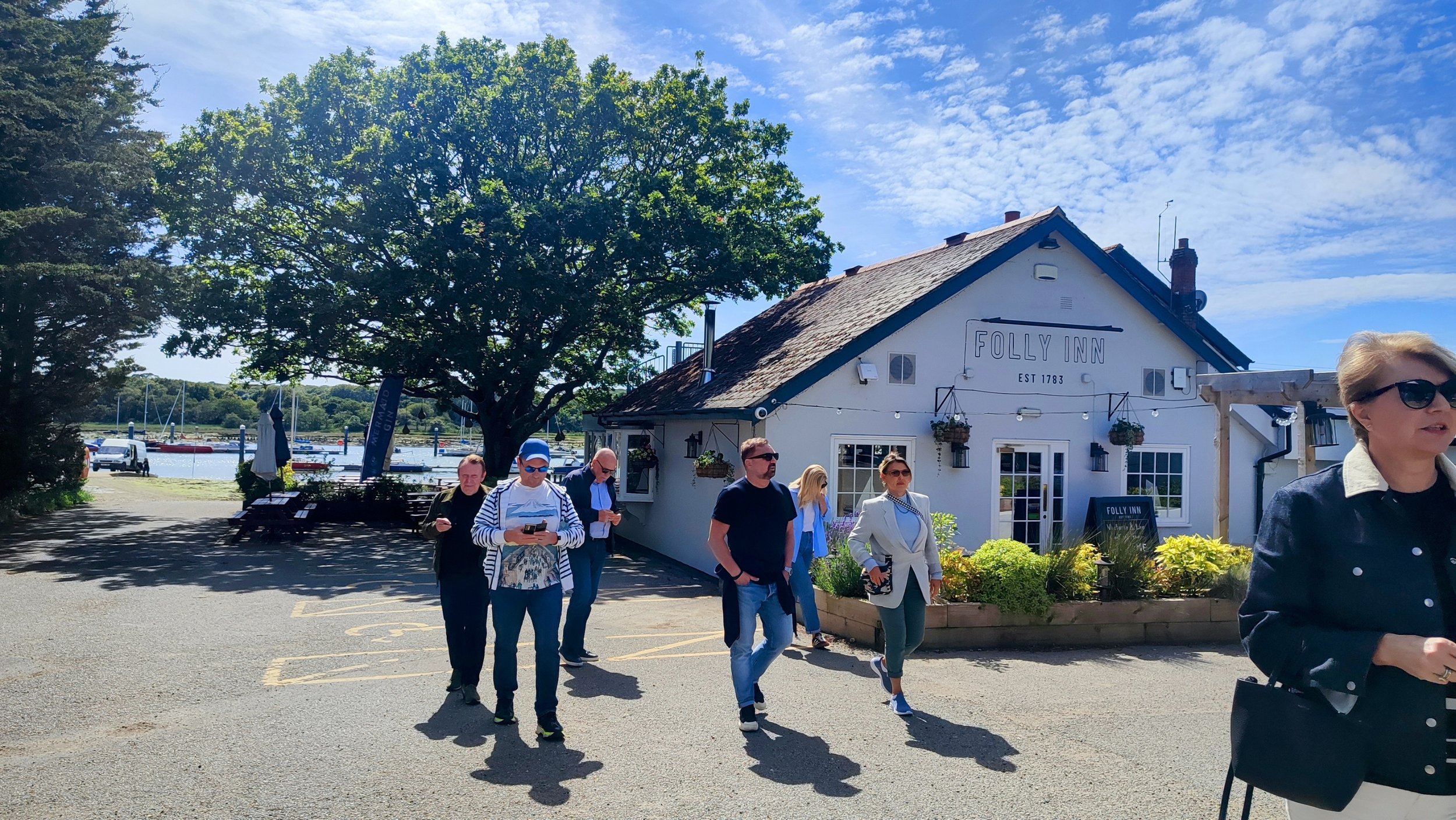 Group of people walking in front of the Folly Inn, a white building with a sign indicating it was established in 1783, near a marina with sailboats docked on a sunny day with partly cloudy sky.