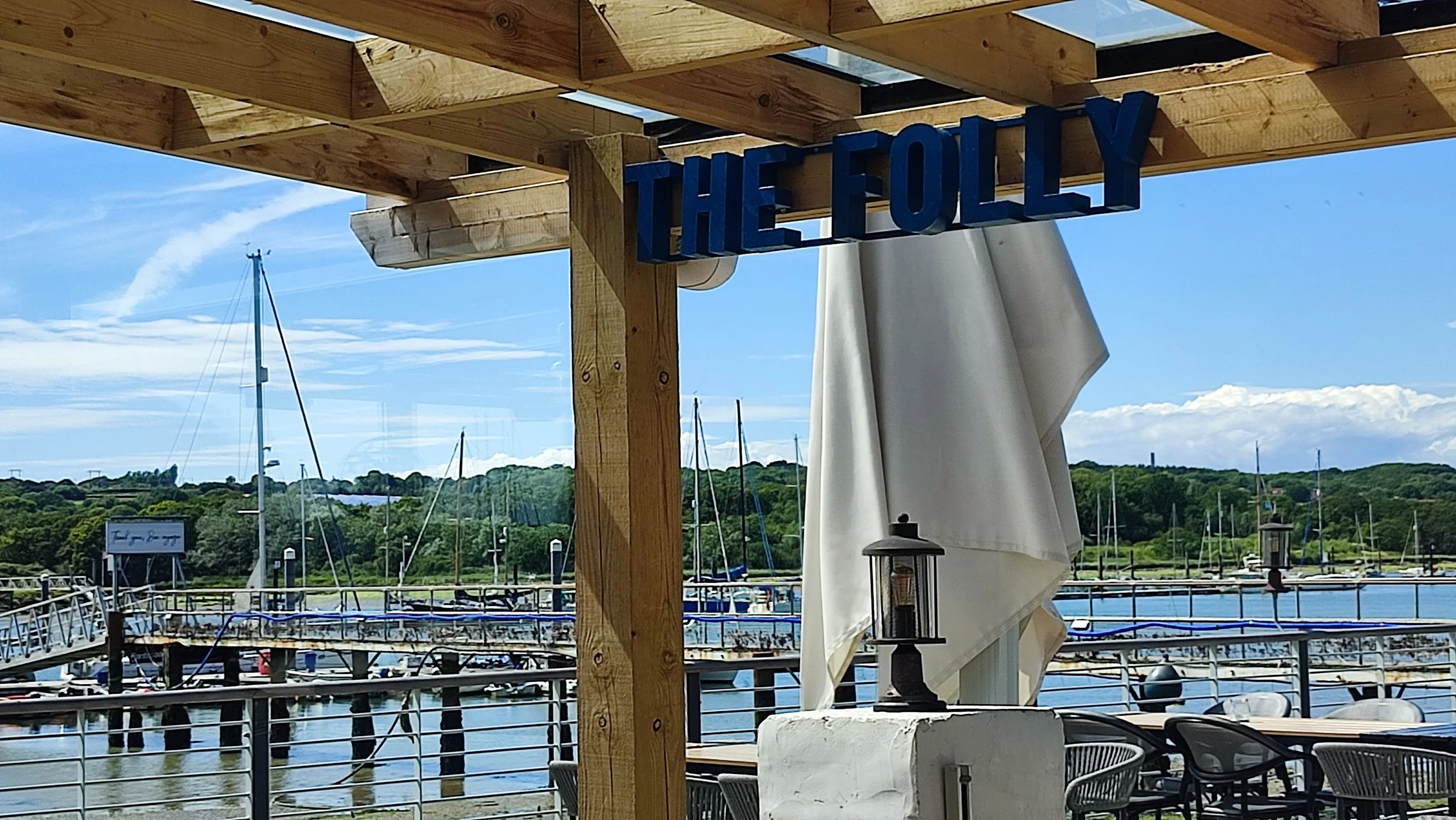 Wooden structure with a sign reading 'THE FOLLY' above a marina with boats and sailboats, outdoor dining tables, and a scenic green hillside in the background on a sunny day.