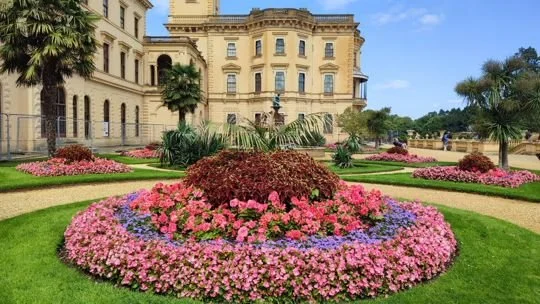 A large historic mansion with a beautifully landscaped garden and flower bed in front, featuring pink, purple, and red flowers, along with palm trees and green grass.