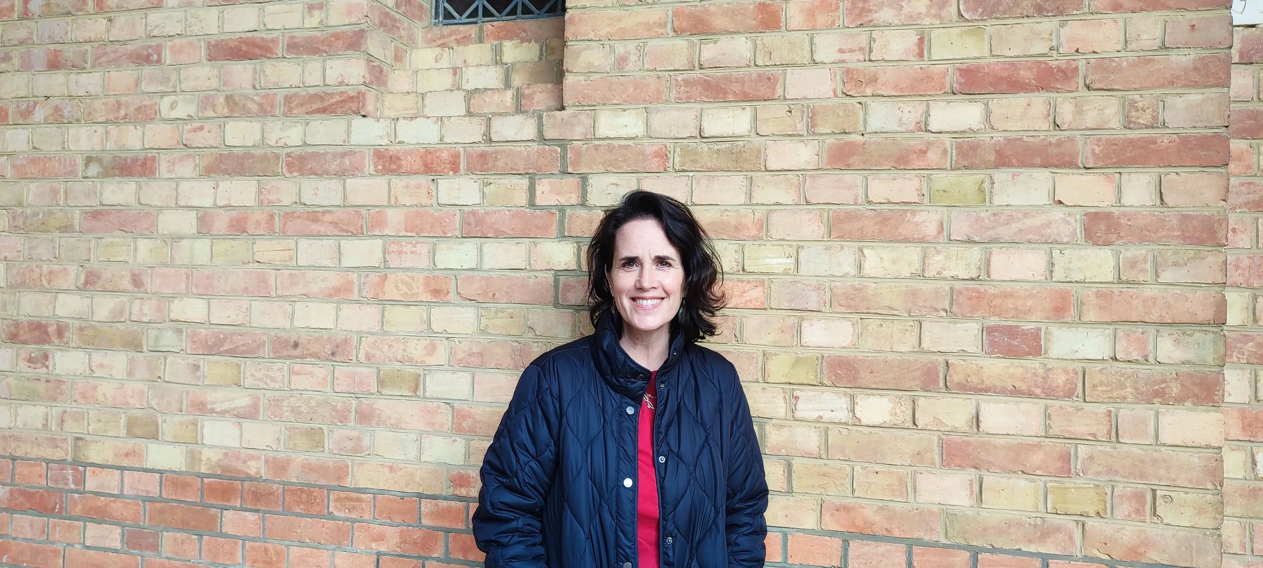 A woman with shoulder-length dark hair wearing a navy blue quilted jacket over a red top, standing in front of a brick wall and smiling.
