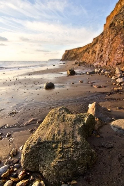 A sandy beach with large rocks and a coastal cliff in the background under a partly cloudy sky.