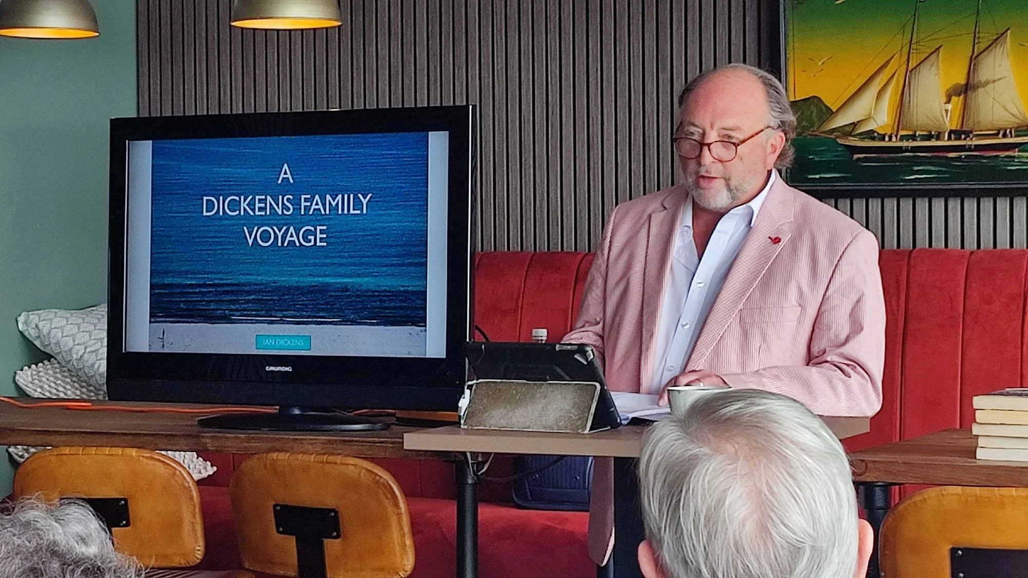 A man with glasses and a beard wearing a pink blazer and white shirt giving a presentation titled 'A Dickens Family Voyage' in a room with a large screen and a painting of a sailing ship on the wall.
