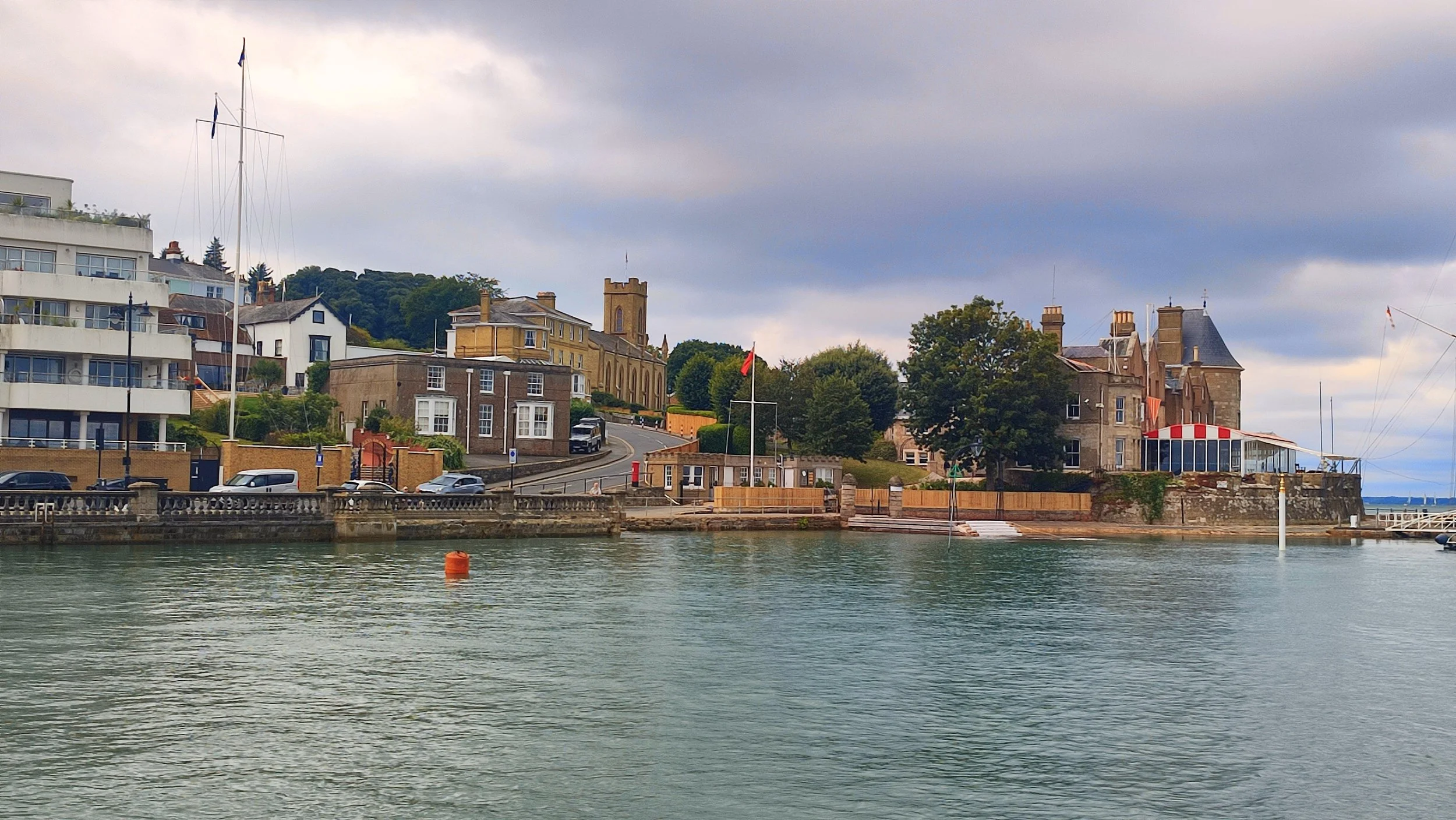 A waterfront scene with calm water in the foreground and a row of buildings along the shoreline. There is a small boat on the right side and a buoy floating in the water. The buildings include residential and possibly commercial structures, some with porches and balconies, and a large tree near the center. A hill with greenery and a tower are visible in the background under a cloudy sky.