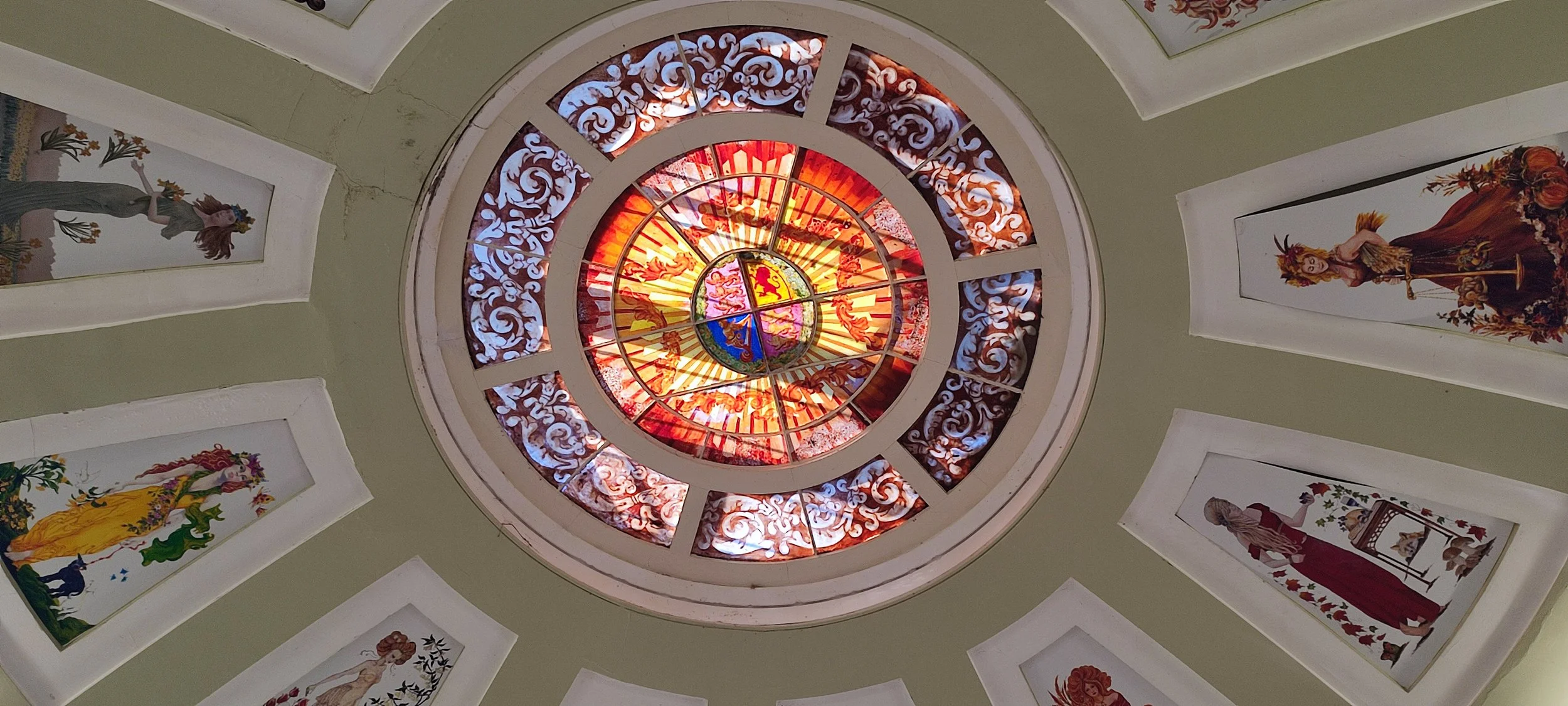 A circular stained glass ceiling with various colors and patterns, surrounded by paintings of women in historical attire on a light green ceiling.