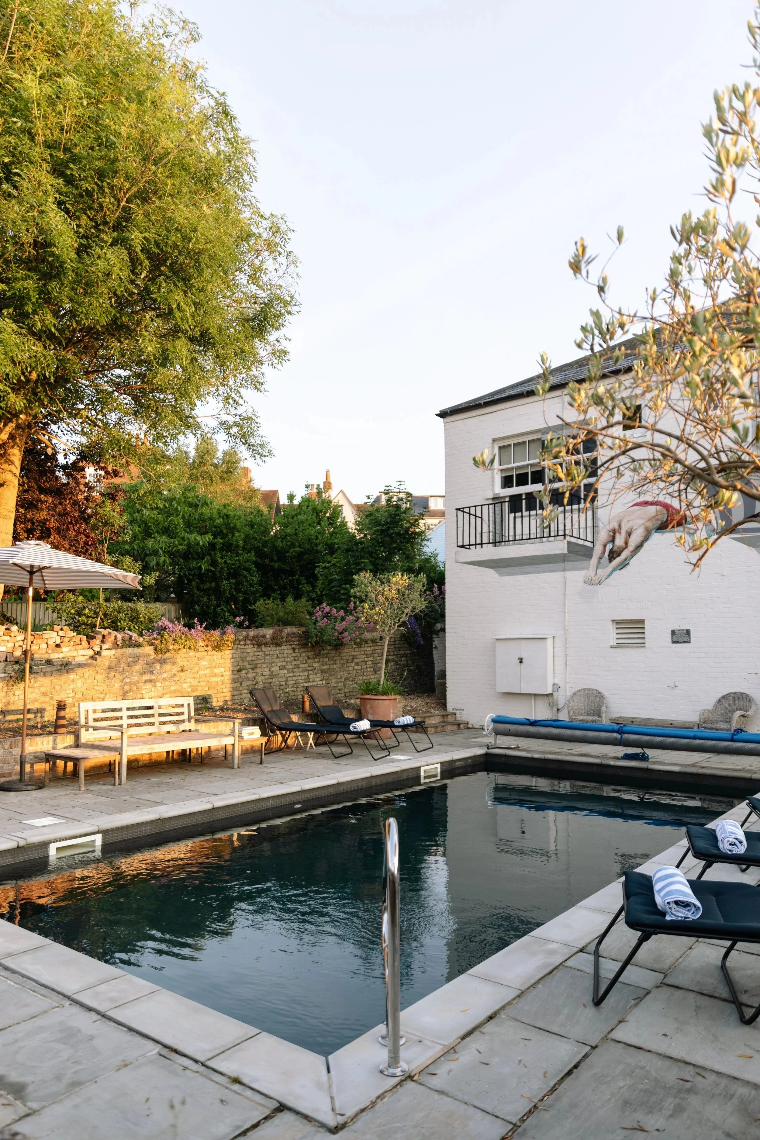 A backyard with a swimming pool, lounge chairs with towels, a large tree, and a white house with a mural of a dog diving from a balcony.
