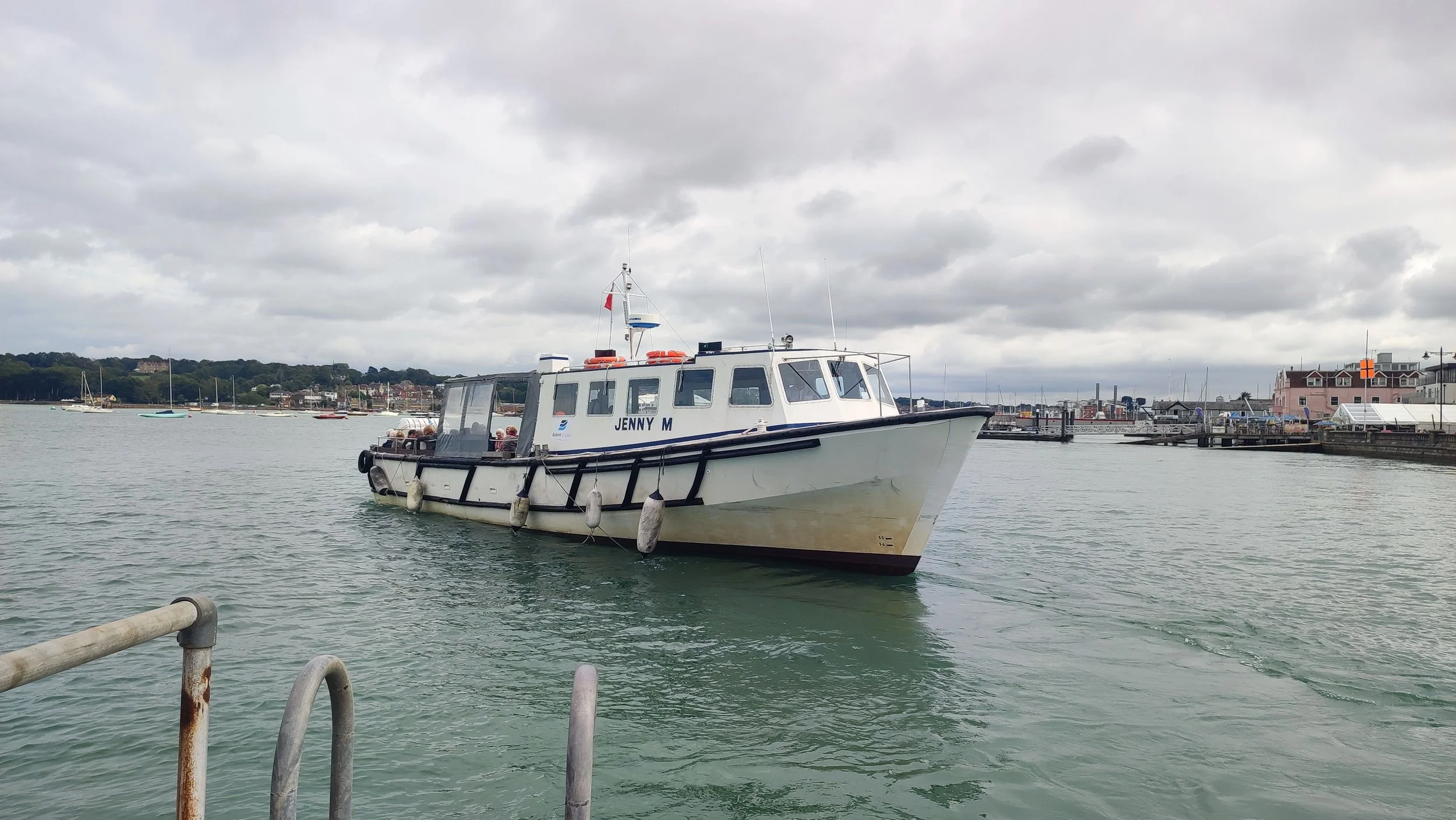 A white boat named Jenny M docked in a harbor with several other boats and buildings visible in the background under cloudy skies.