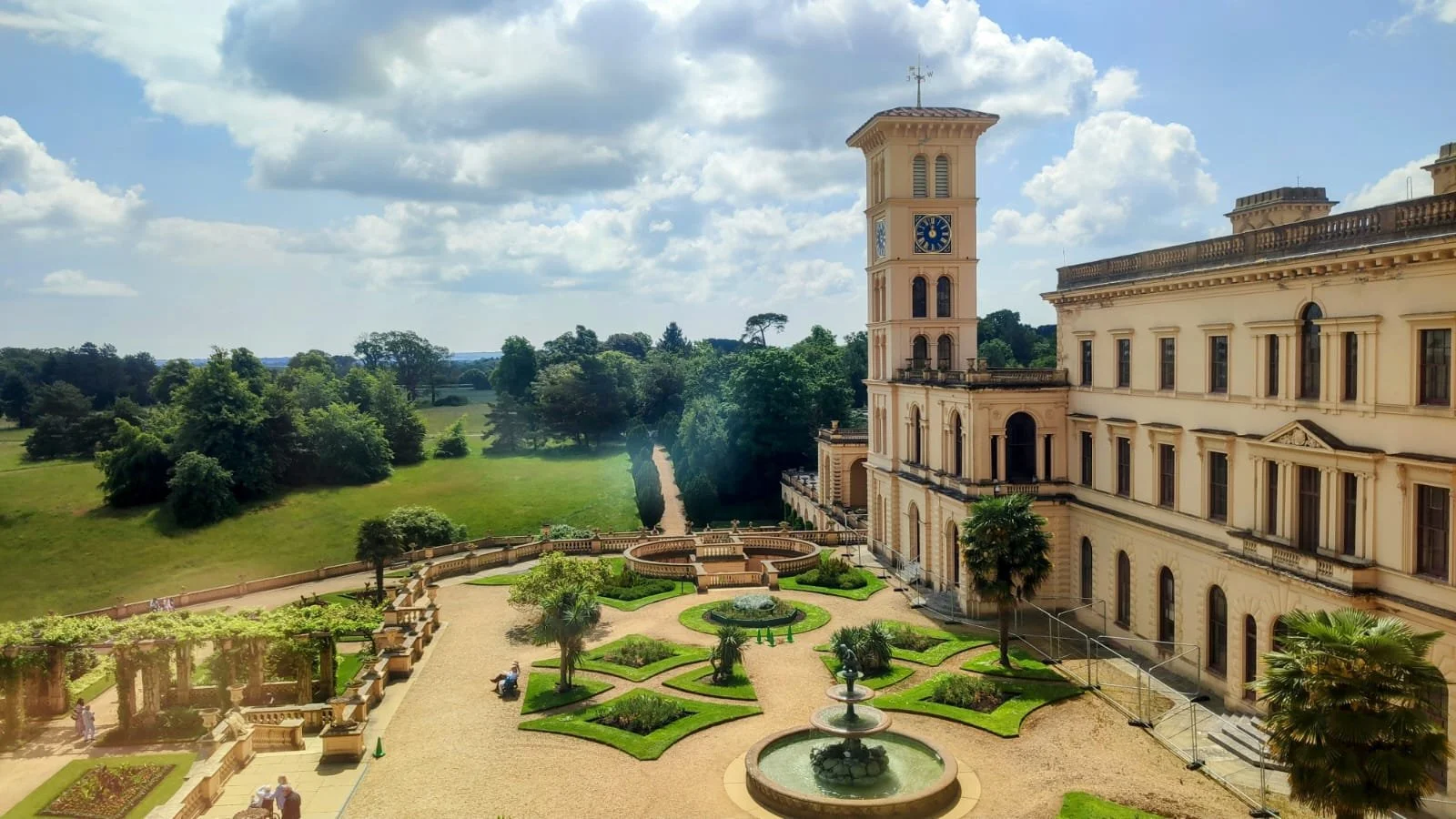 A historic building with a clock tower, surrounded by a well-kept garden with fountains, trees, and a gravel path, under a partly cloudy sky.