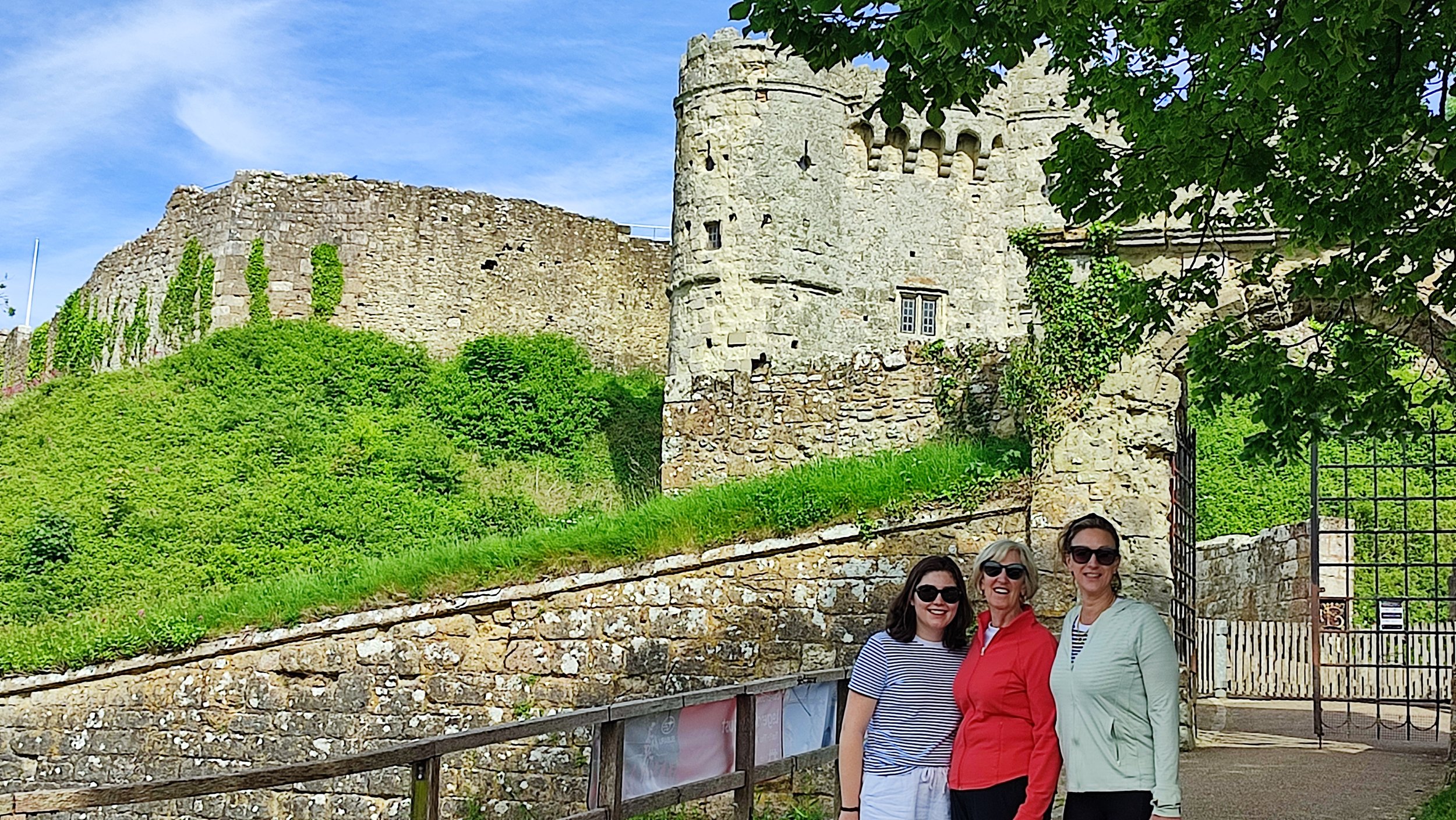 Three women standing in front of a castle with stone walls and a tower, under green trees and a blue sky.