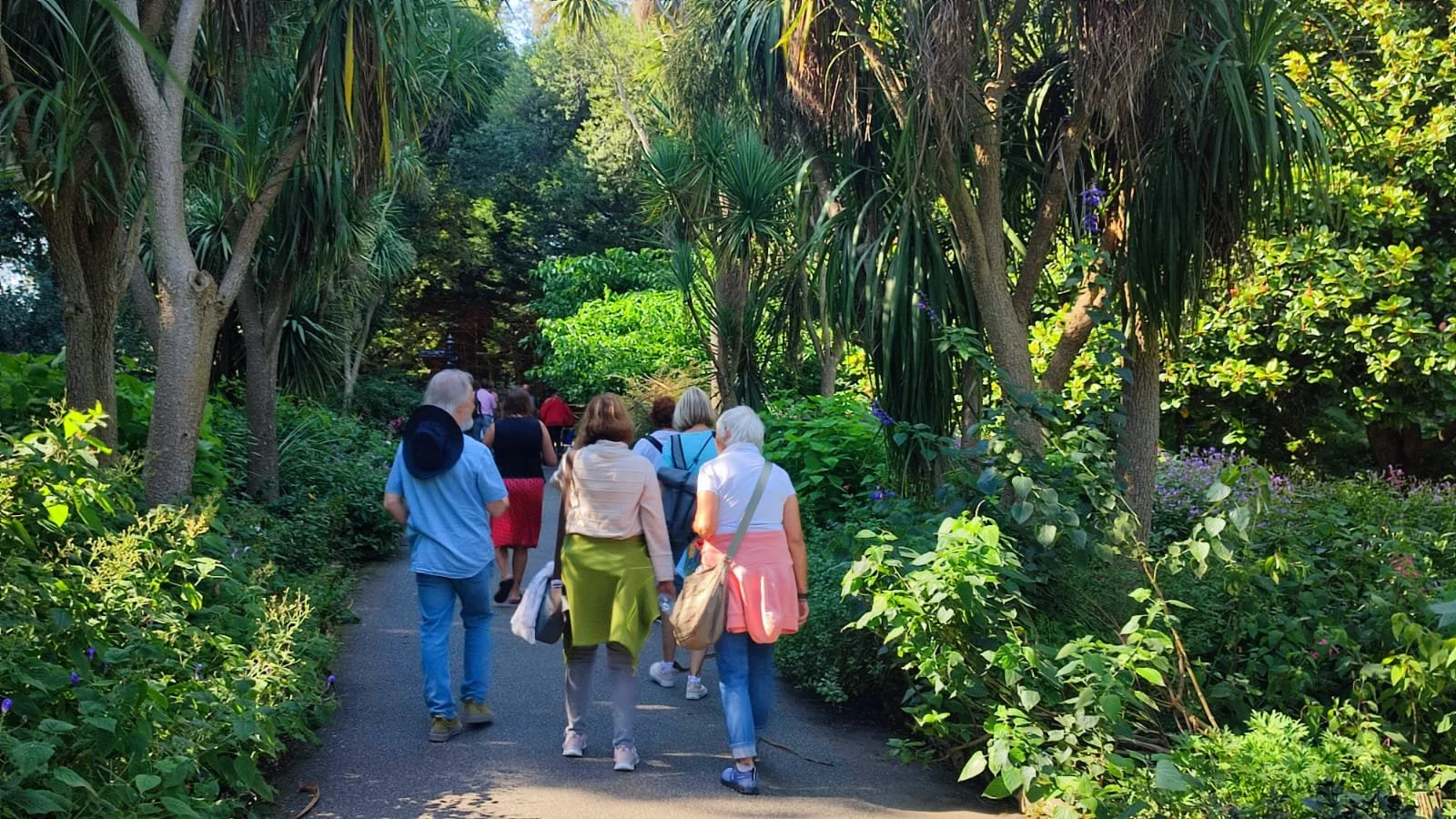 Group of people walking along a lush, green garden pathway surrounded by dense trees and plants.