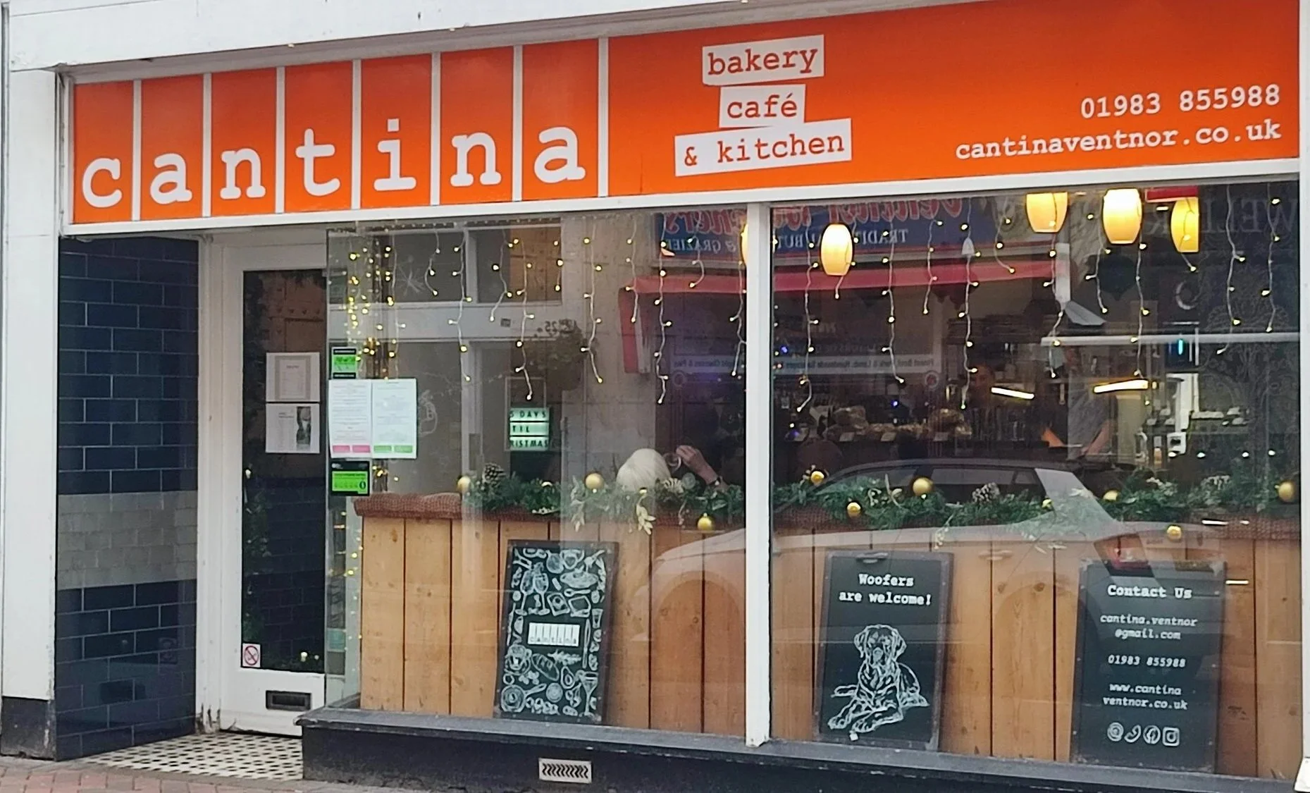 Storefront with orange sign reading 'cantara bakery cafe & kitchen' and contact information, decorated with Christmas ornaments and fairy lights, with visible interior of a bakery and customers inside.