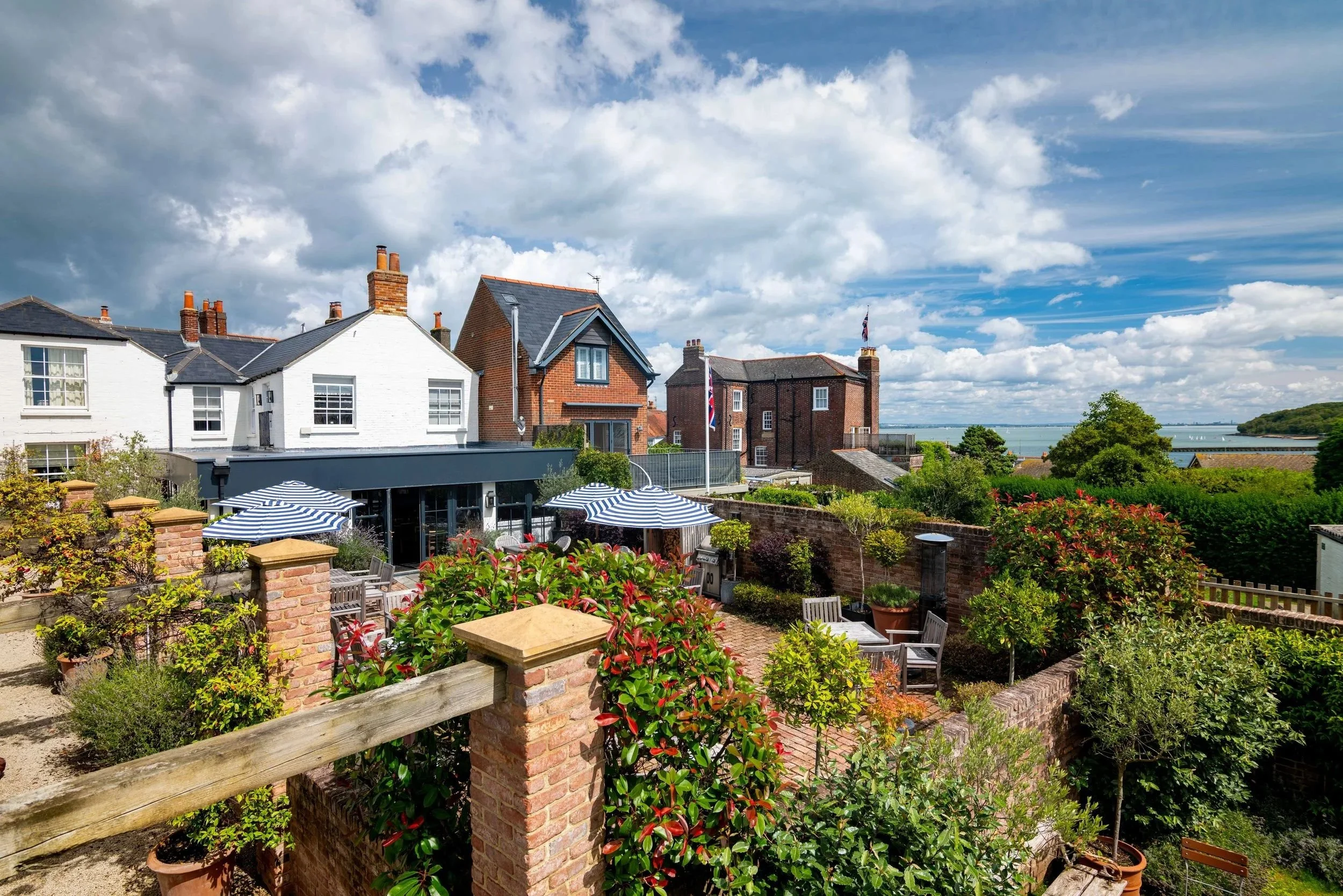 A picturesque view of a garden with outdoor seating area, umbrellas, and lush greenery, overlooking a neighborhood with houses and a body of water in the background under a partly cloudy sky.
