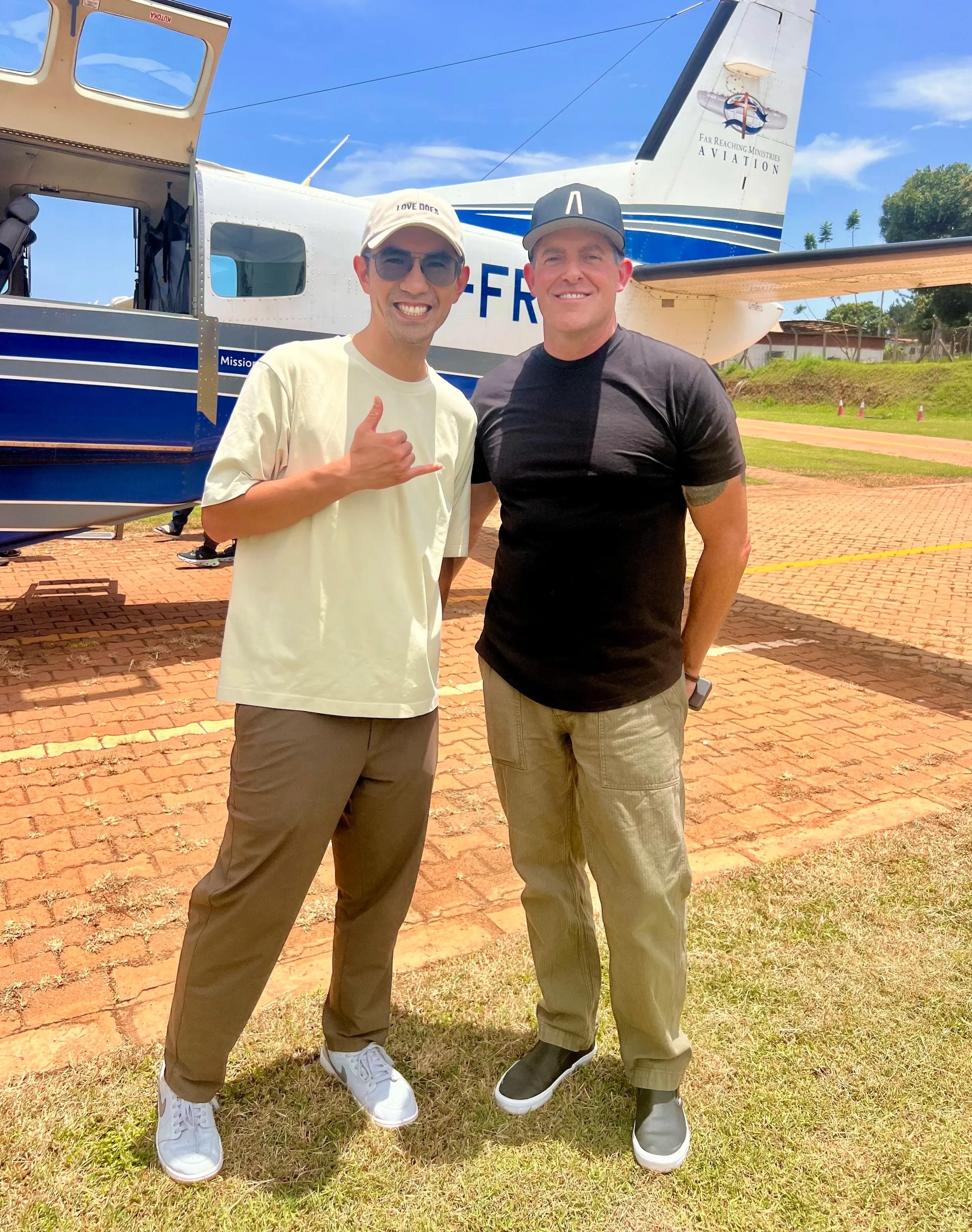 Two men standing outdoors in front of a small airplane, smiling at the camera. One is making a hand gesture, wearing sunglasses, a baseball cap, a light-colored t-shirt, and khaki pants. The other is wearing a dark t-shirt, a baseball cap, and beige pants. The airplane behind them is white with blue accents and has the words 'FAR REACHING MINISTRIES AVIATION' on the tail.