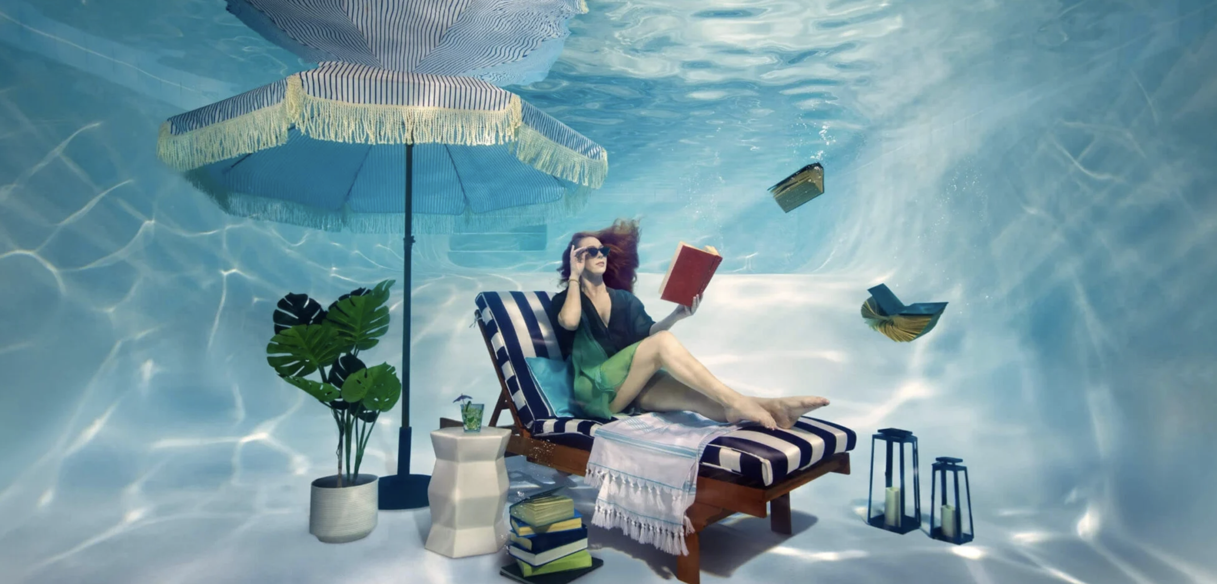 Woman relaxing on a poolside lounge chair underwater, reading a book, with an umbrella, potted plants, and floating papers around her.