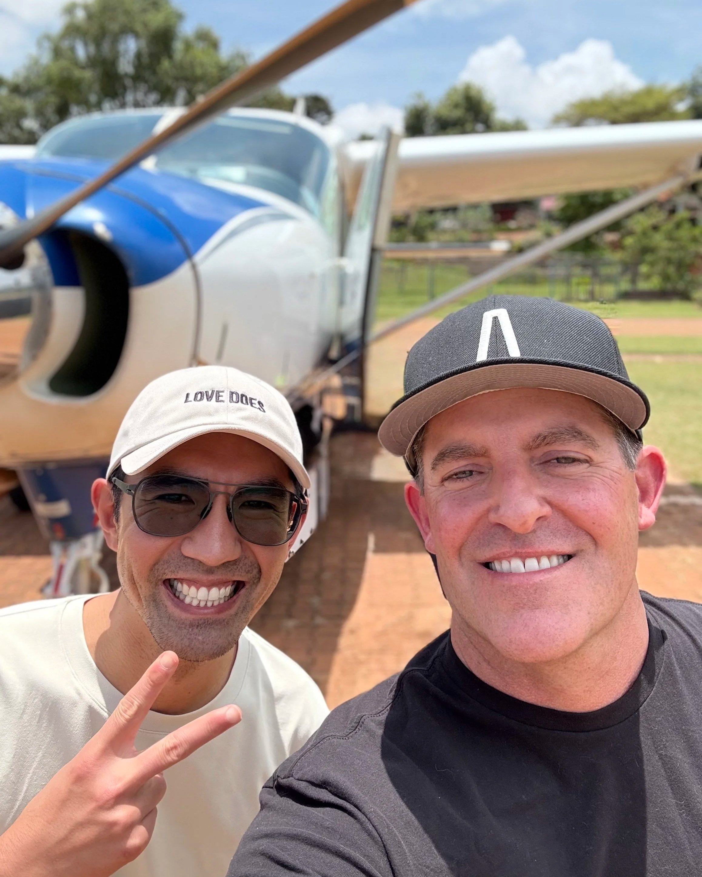 Two smiling men taking a selfie in front of a vintage airplane with blue, white, and wood finish at an outdoor airfield or park.