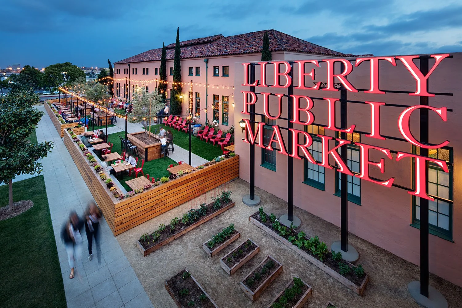 Outdoor seating area at Liberty Public Market during evening with string lights, people dining, and a large pink neon sign on the building.