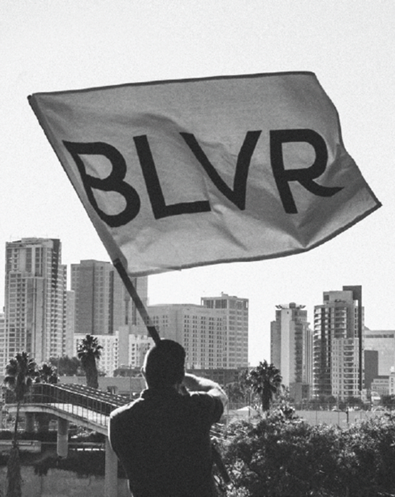A person holding a large flag with the word 'BLVR' on it against a cityscape background with tall buildings and palm trees.