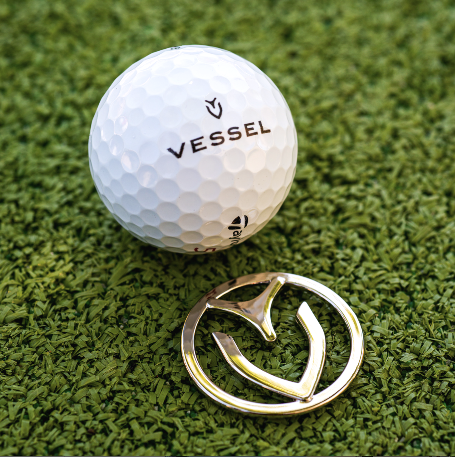 A close-up of a golf ball with the brand name 'Vessel' and a logo, placed on artificial green grass, with a metal golf tee lying next to it.