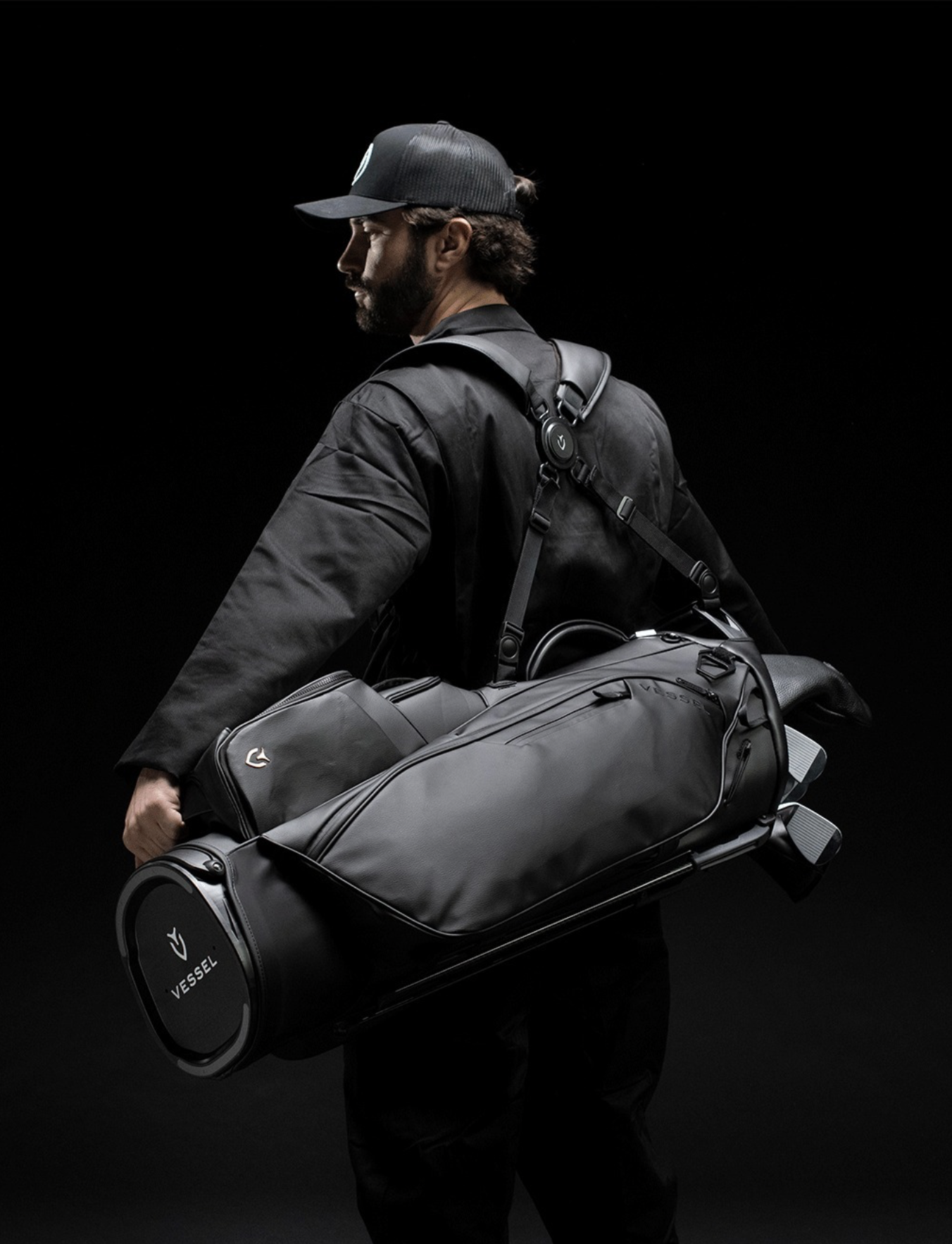 A man wearing a black cap and black jacket, carrying a large black golf bag with a stand against a black background.