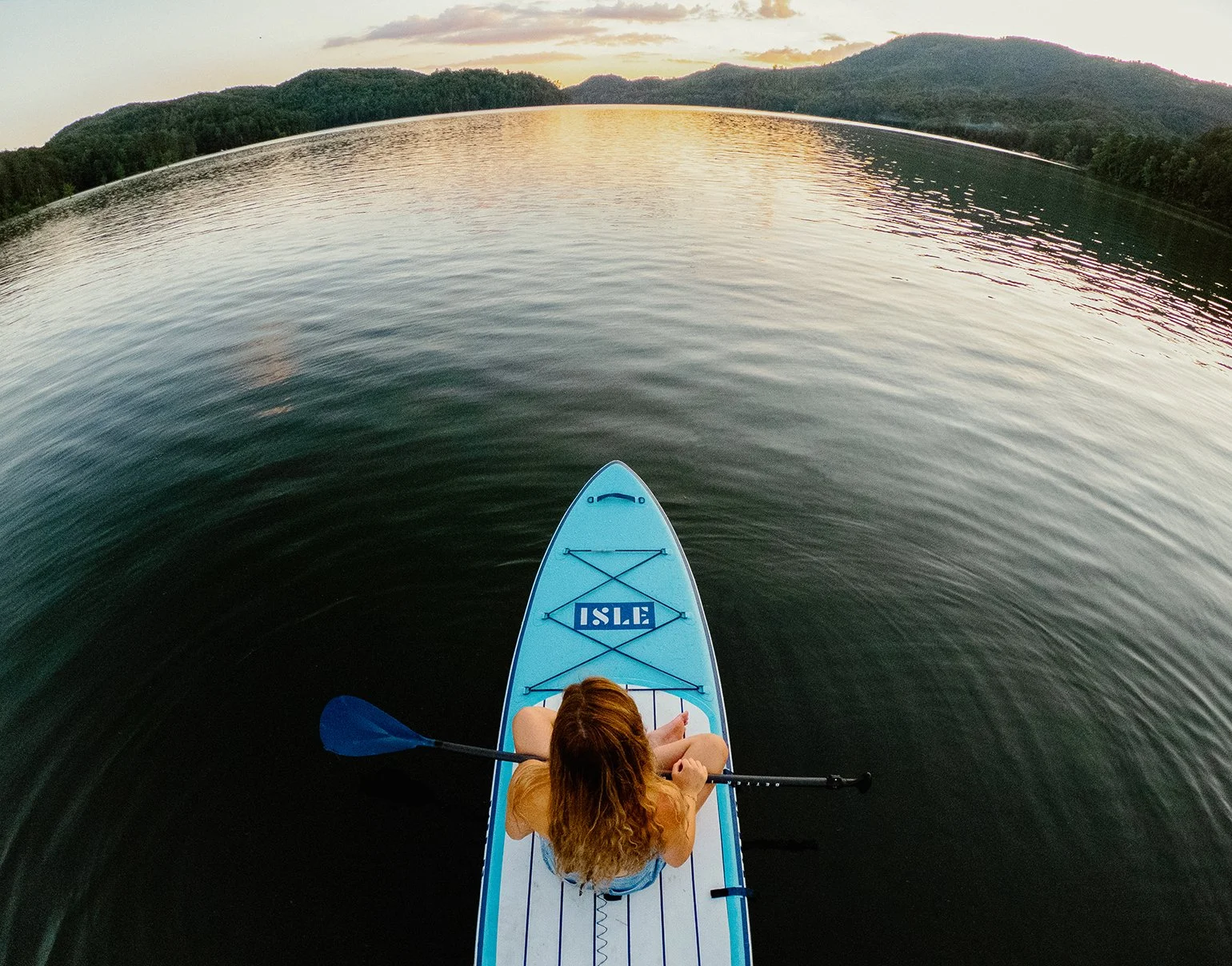 A person with long hair paddling a blue stand-up paddleboard on a calm body of water during sunset, surrounded by green hills.