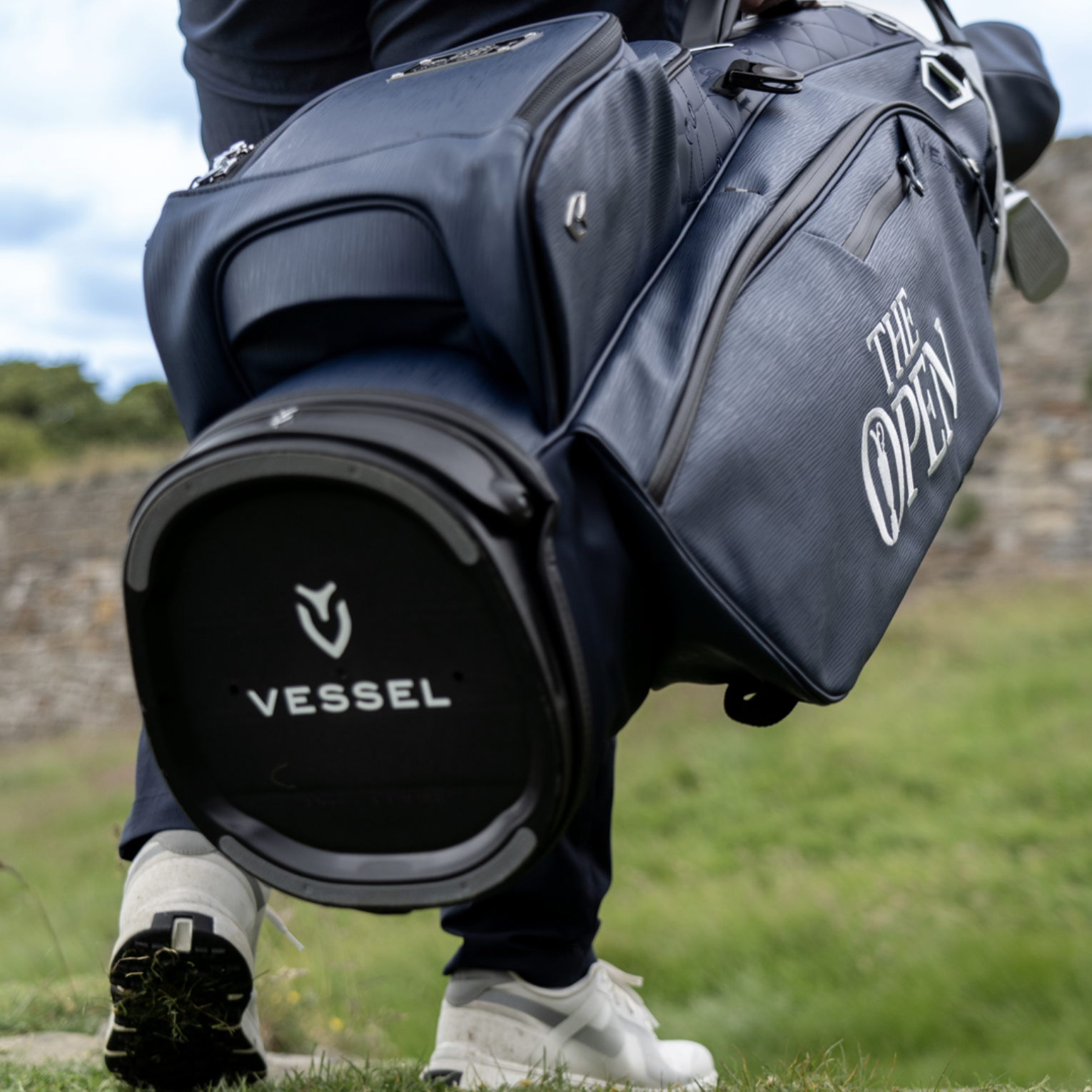 Close-up of a person holding a VESSEL golf bag with 'THE OPEN' logo, outdoors with grass and trees in the background.