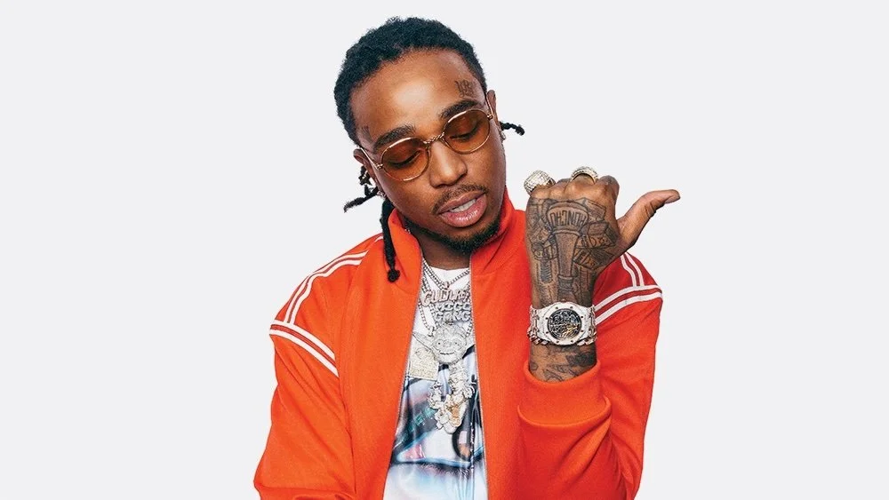 A portrait of a man with dreadlocks wearing sunglasses, a red sporty jacket, various jewelry including a necklace and watch, and showing tattoos, posing with one hand raised under his chin against a plain light background.
