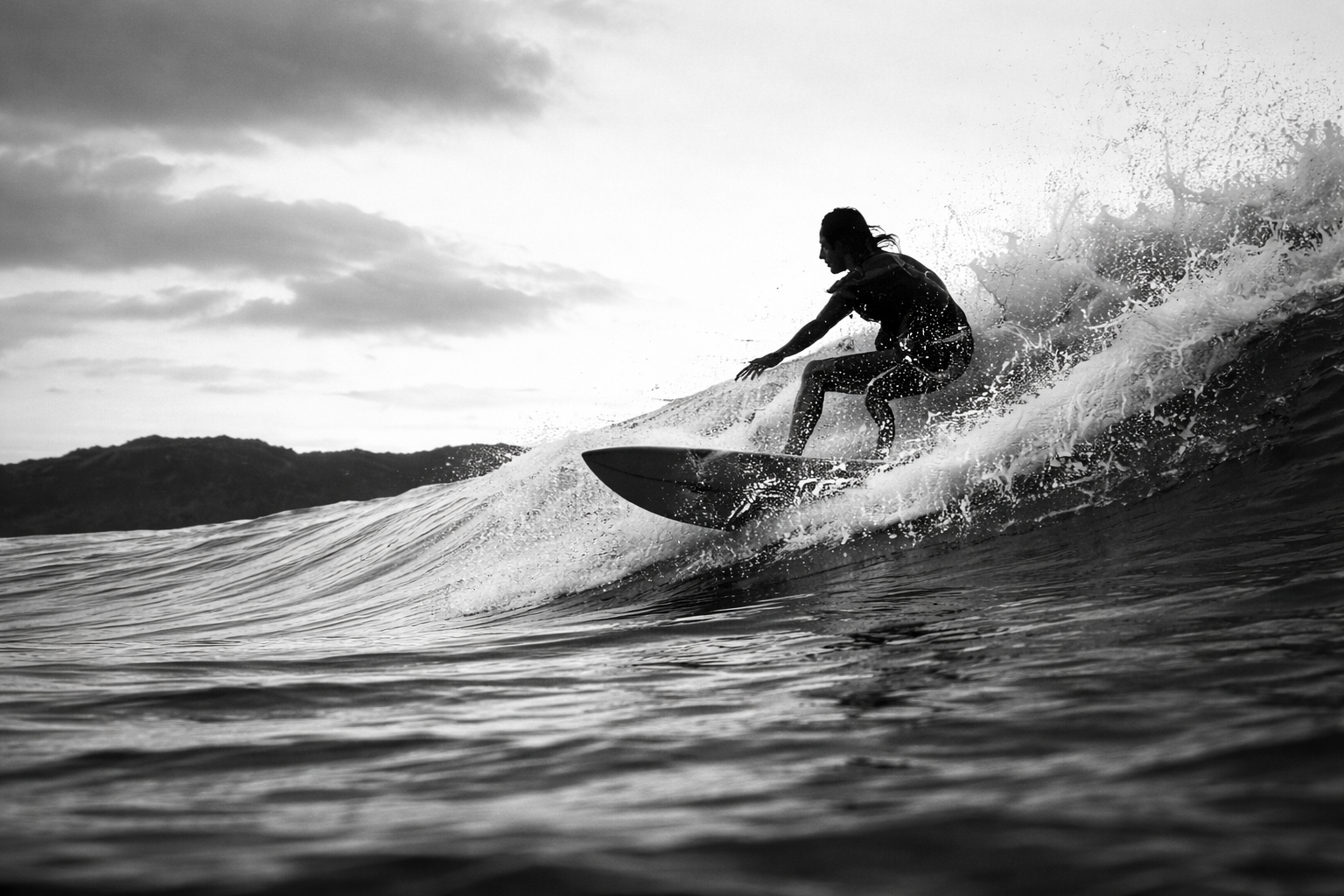 A person surfing on a wave during cloudy weather, captured in black and white. Assistant is riding a surfboard on the crest of a wave.