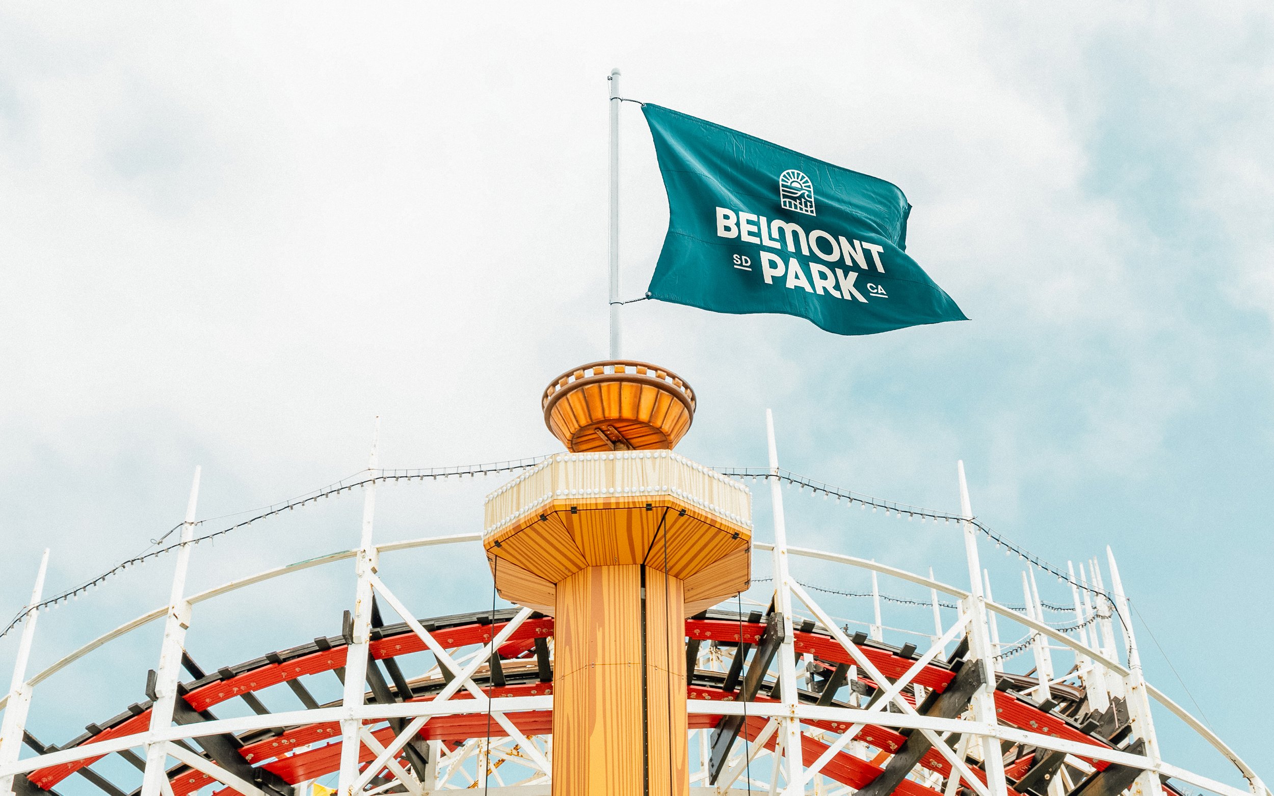 View of a Ferris wheel at Belmont Park with a flag reading 'Belmont Park' flying at the top, against a cloudy sky.