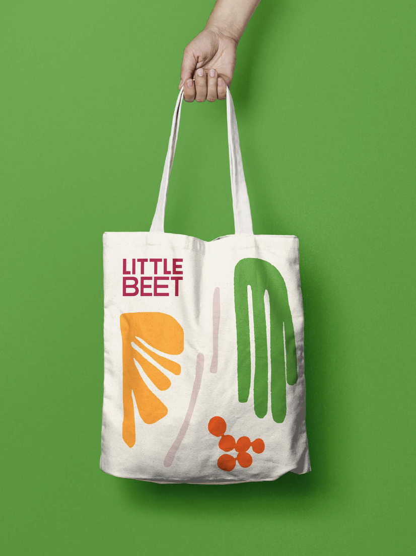 A hand holding a white tote bag featuring colorful abstract vegetable illustrations and the words "LITTLE BEET" on a green background.