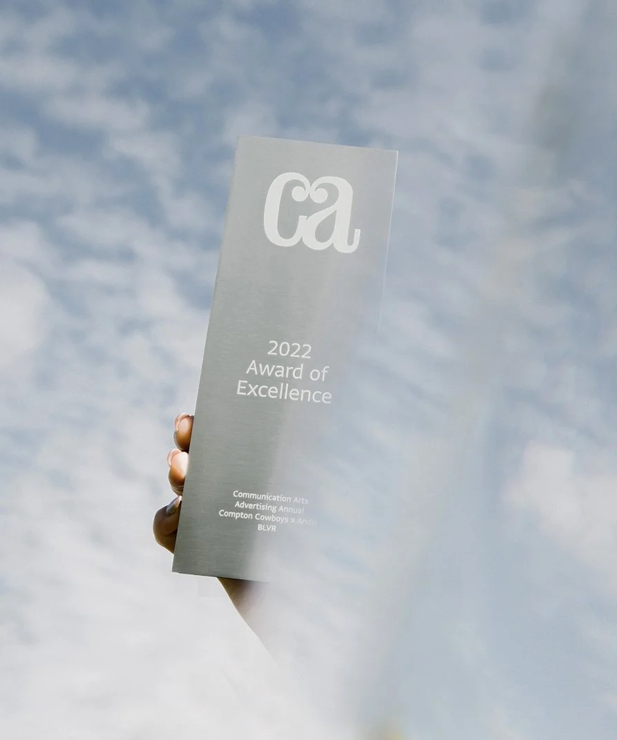 Hand holding a silver award plaque with sky background, labeled '2022 Award of Excellence' from CA for Communication Arts Advertising Annual.