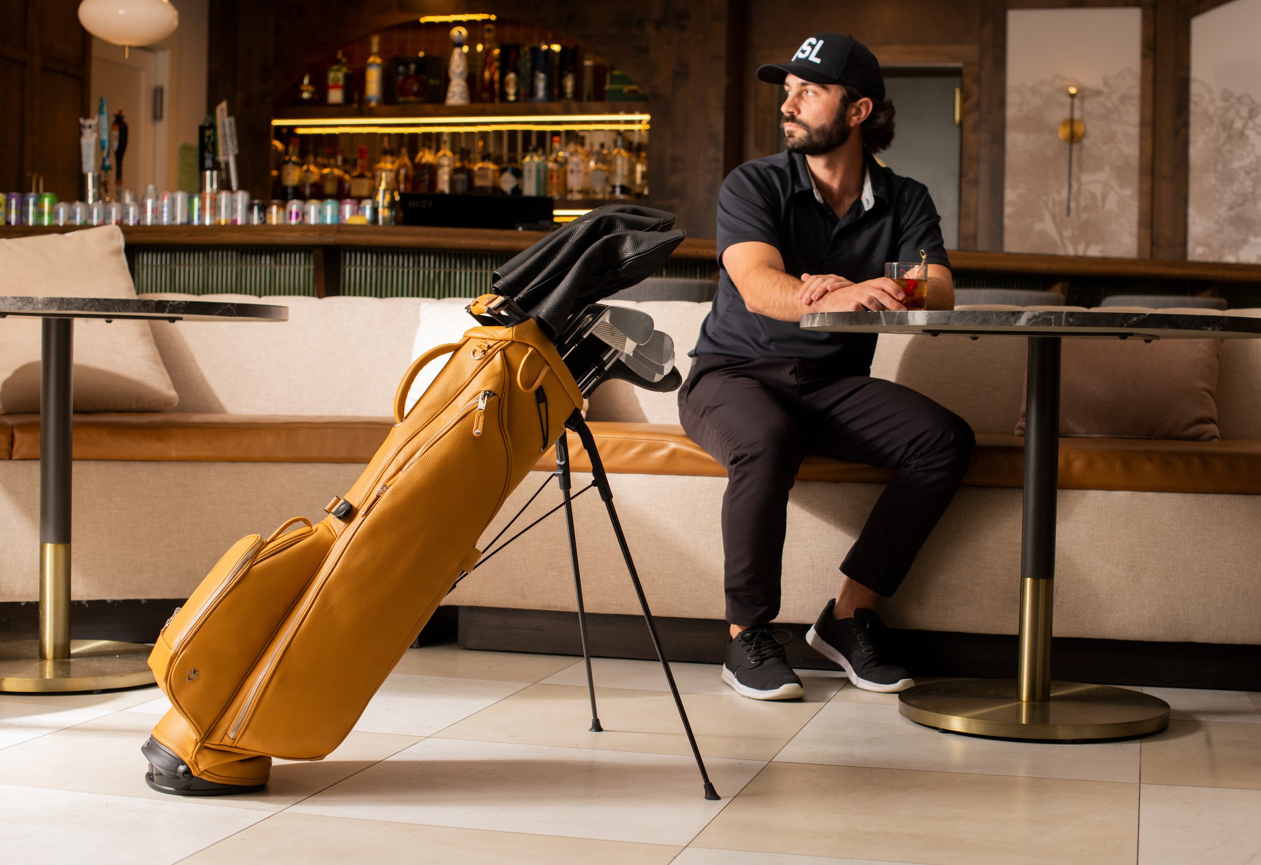 A man sitting in a bar or lounge area with a glass of drink, a yellow golf bag with clubs leaning against a sofa, and a bar in the background.