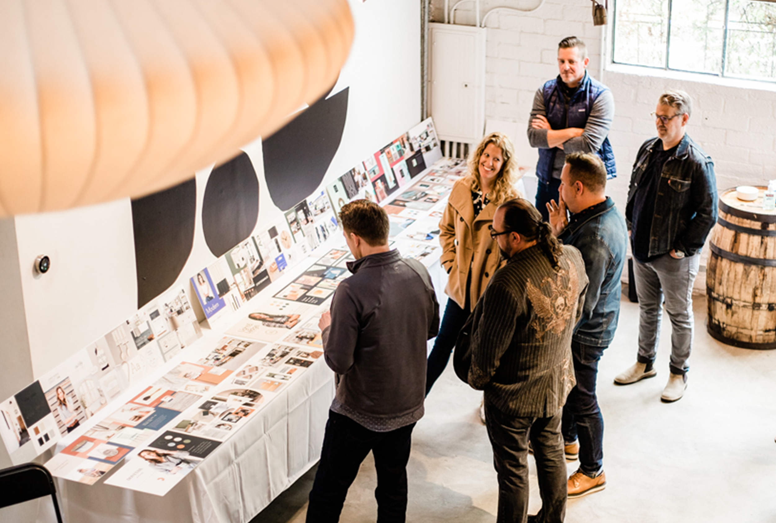 Group of people viewing a presentation or art display on a table in a well-lit room with white brick walls and large windows.