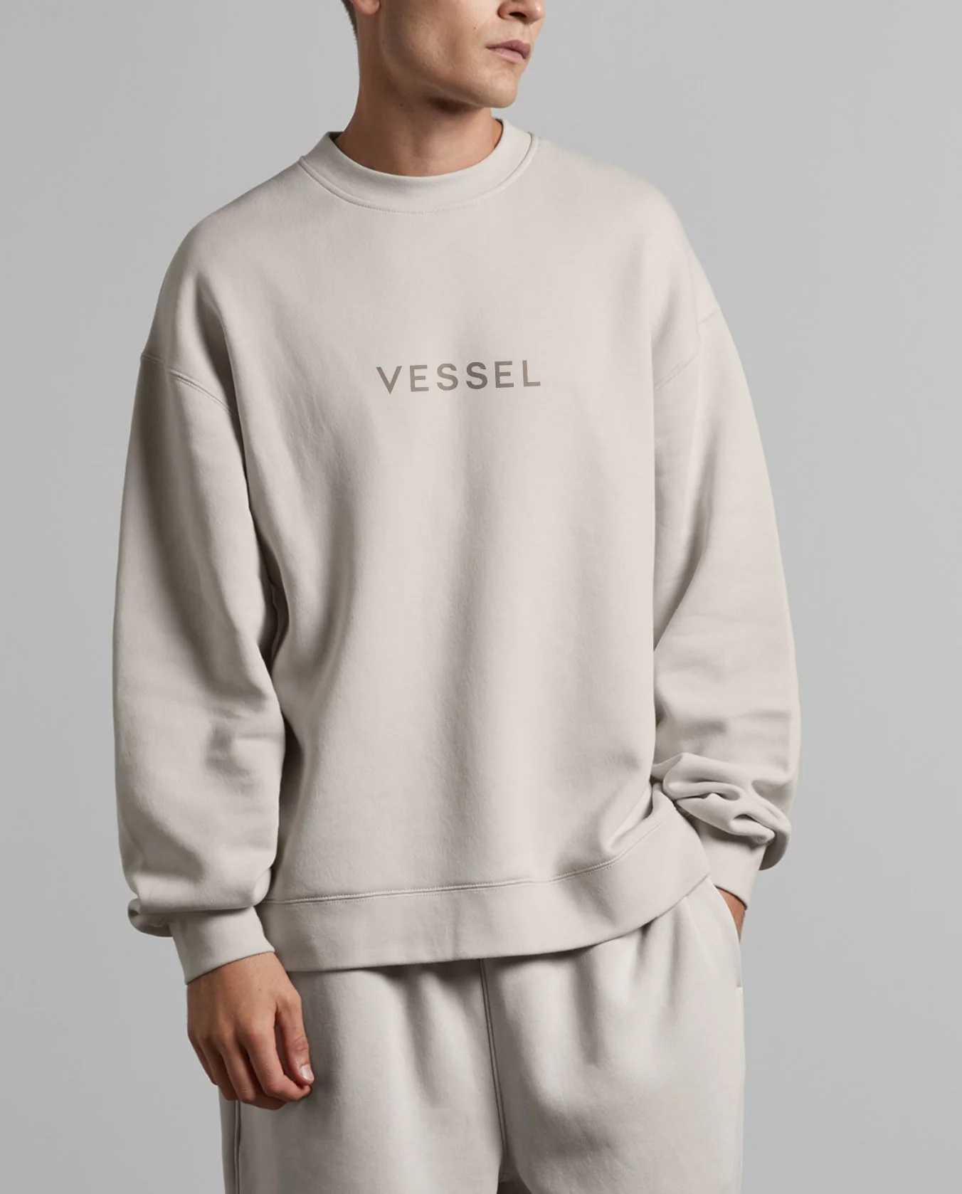 A person in a beige sweatshirt with the word 'VESSEL' written across the chest, standing against a plain gray background.