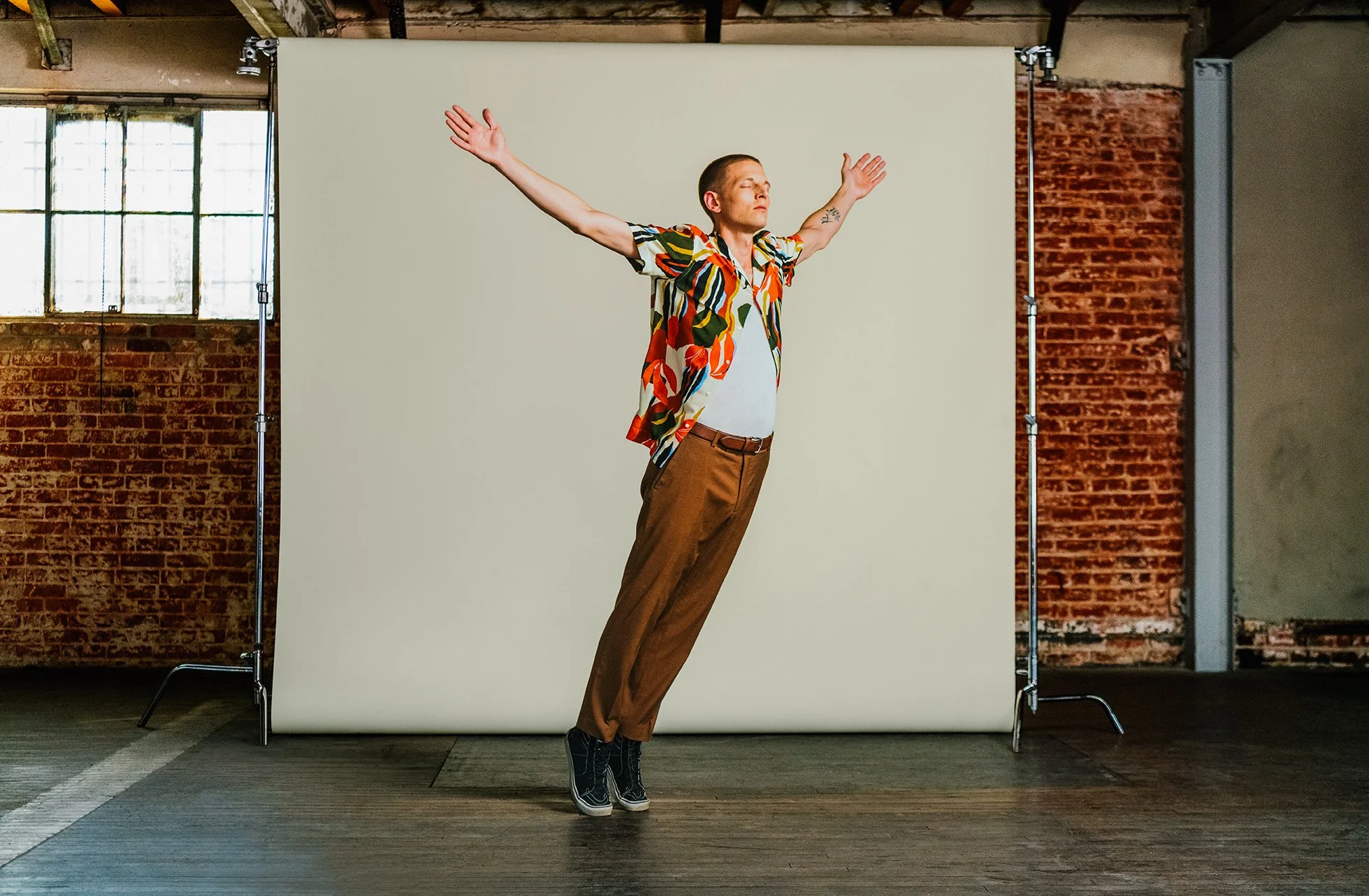Man with arms outstretched, standing in front of a white backdrop in an industrial-style room with large windows and exposed brick walls.