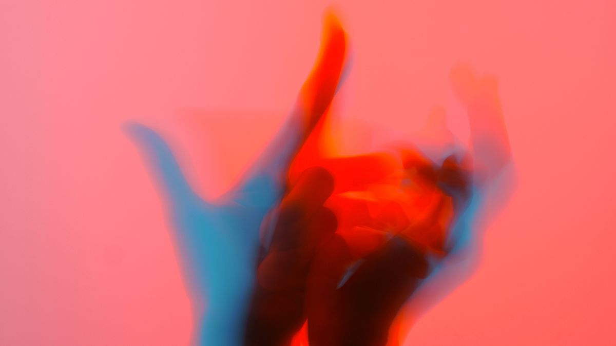 Blurry, colorful hand gesture with red, blue, orange, and black hues on a pink background.