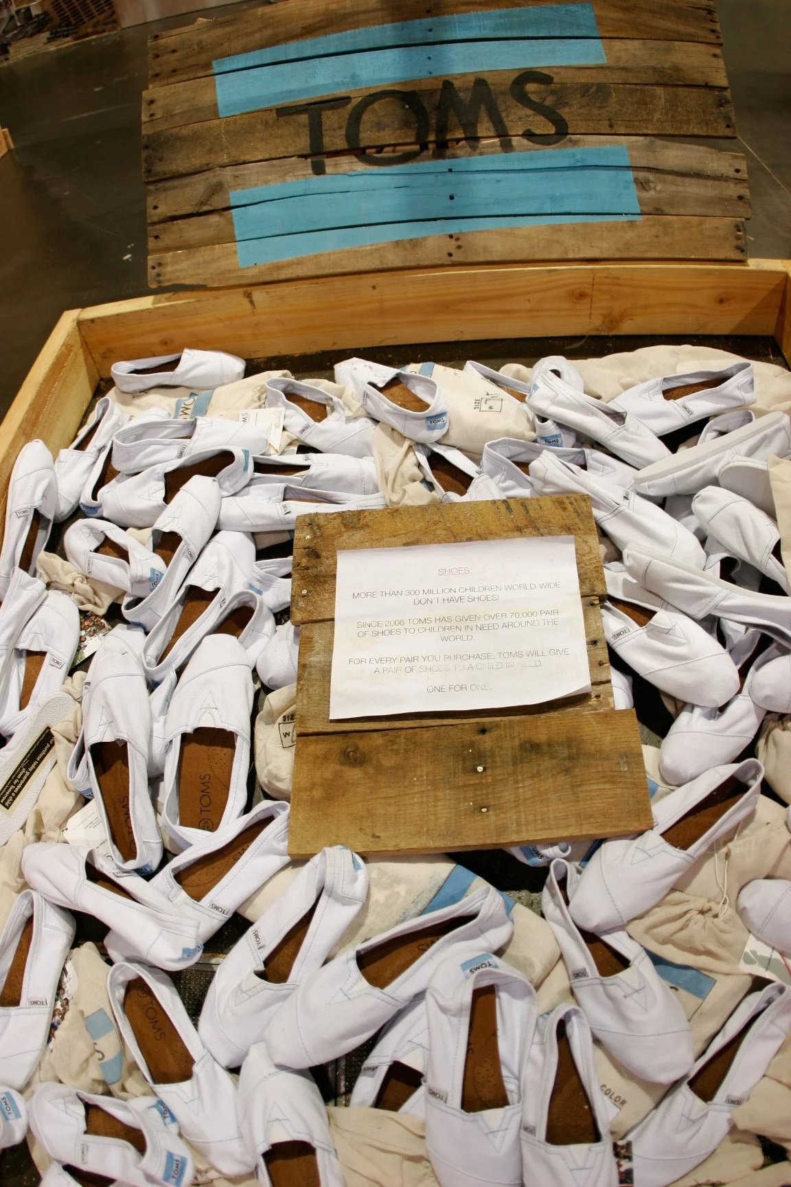 A large pile of white children's shoes inside a wooden container with a sign explaining the TOMS philanthropic initiative, which donates a pair of shoes for every pair purchased.