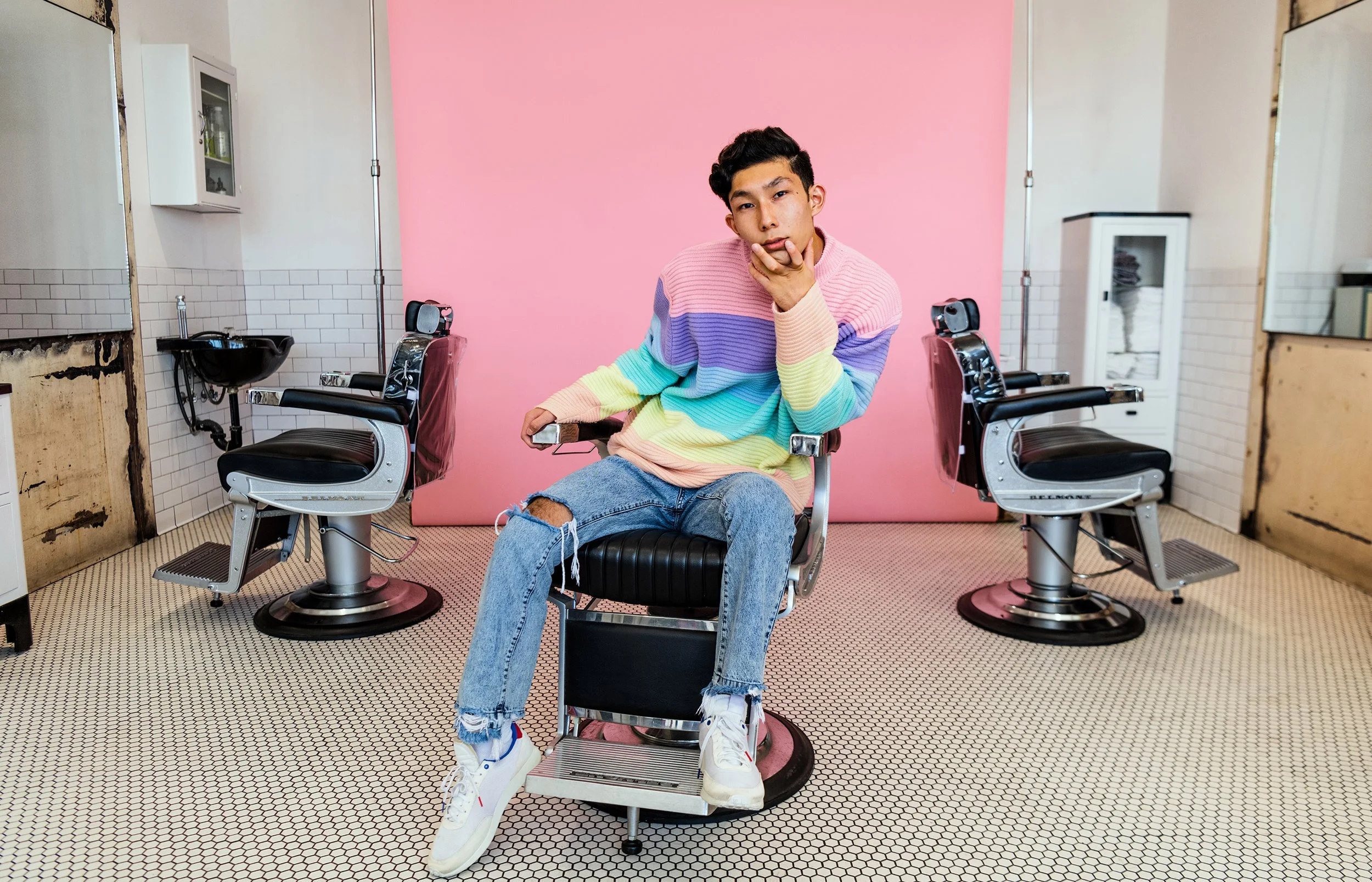 A young man wearing a colorful striped sweater and ripped jeans sits on a vintage barber chair in a barber shop with two empty chairs and a pink wall in the background.