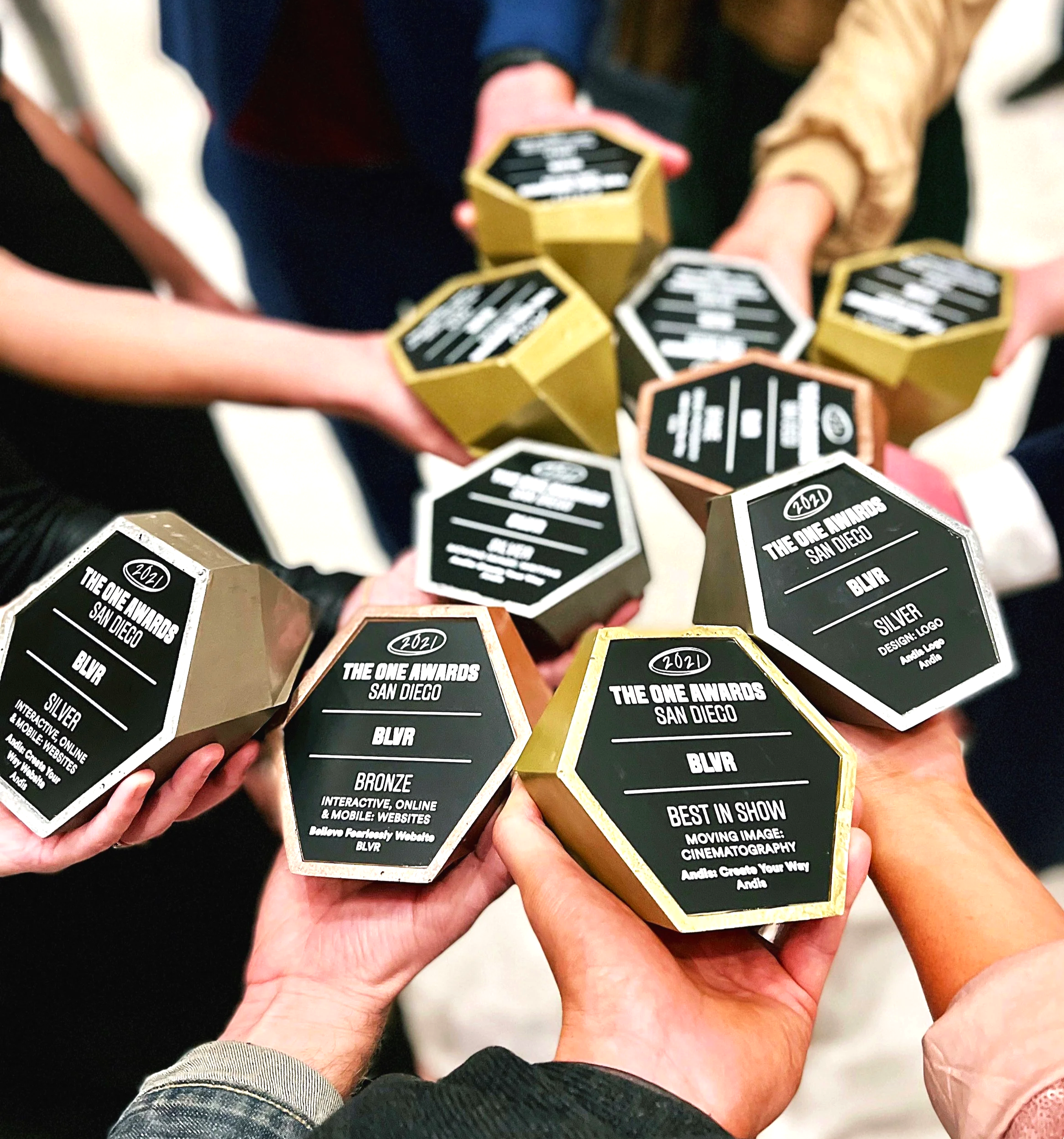 Multiple people holding awards with black plaques and gold or pink borders at 'The One Awards 2021 San Diego' ceremony.